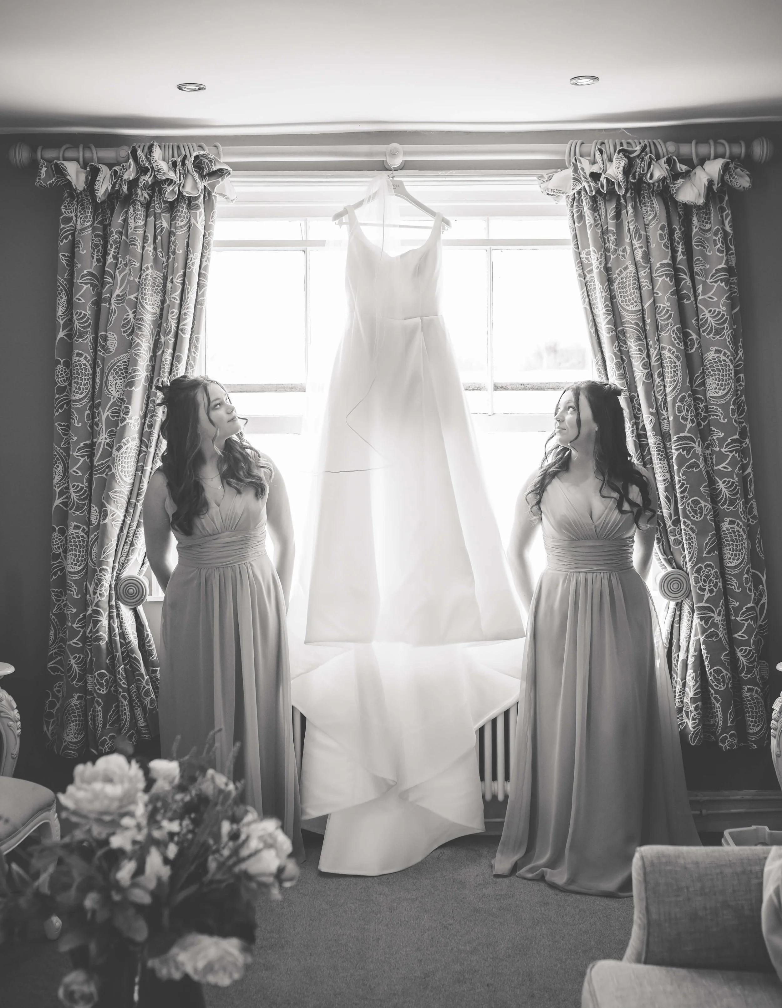 Two women in matching dresses standing on either side of a window with a hanging wedding dress in the middle, all backlit by natural light.