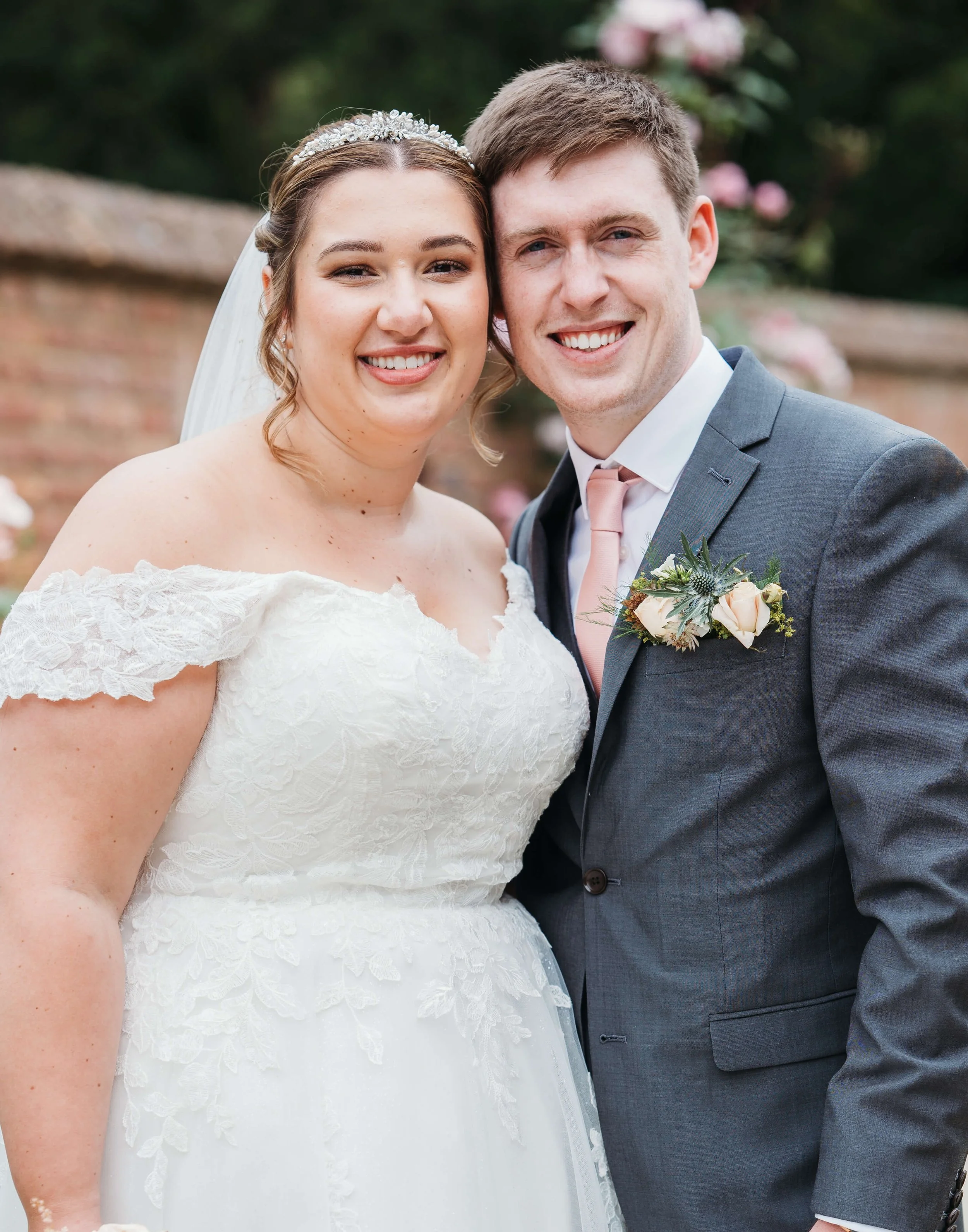 A bride and groom smiling at their wedding, standing close together outdoors with a brick wall and flowers in the background.