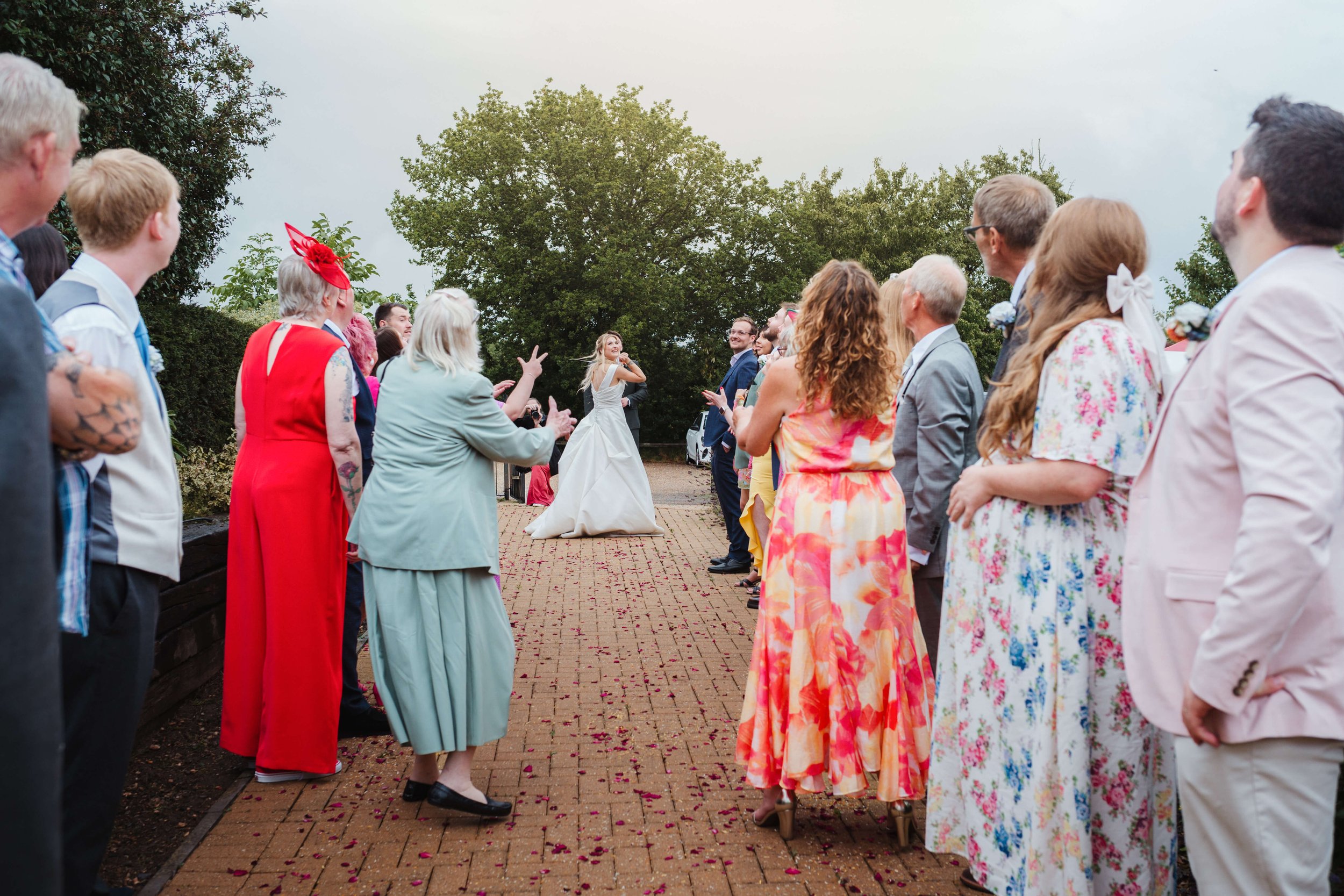 A wedding ceremony with guests forming a semi-circle around the bride, who is dancing and smiling, outdoors with trees in the background.