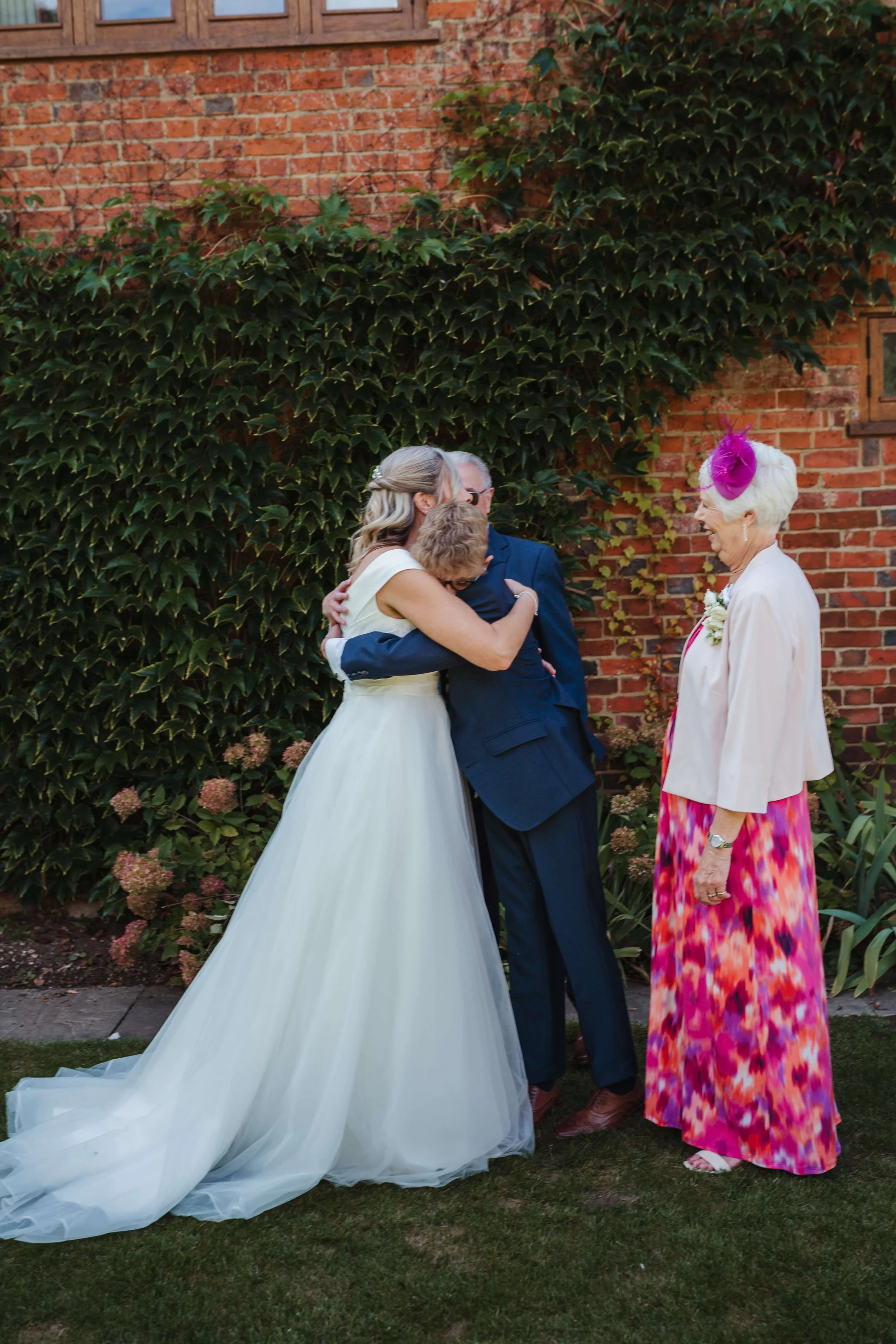 A bride in a white wedding gown hug a man in a suit while an older woman in a colorful dress and pink hat looks on in a garden with a brick wall and greenery.