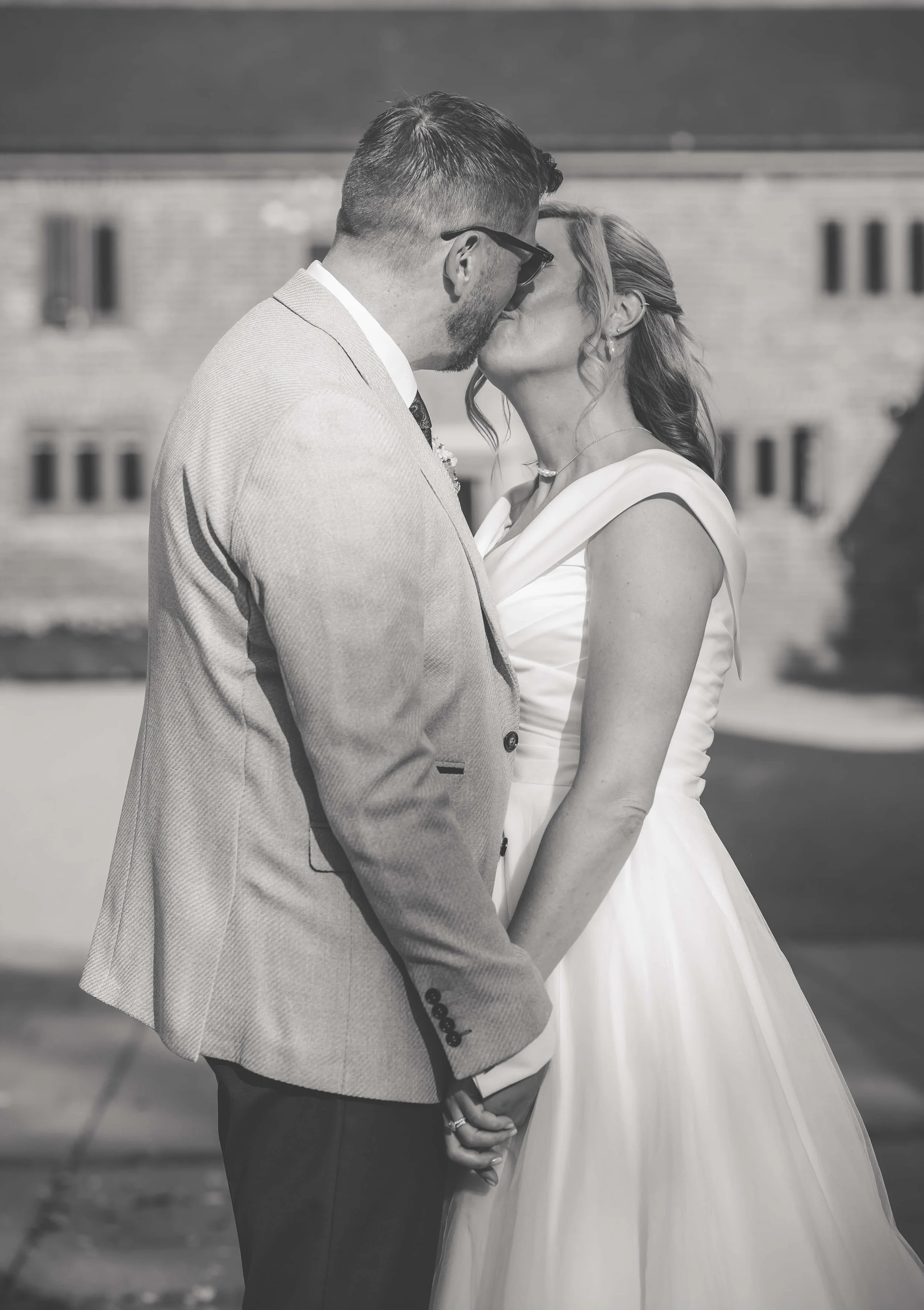 A black-and-white photo of a man and woman kissing outdoors, with the man in a suit and the woman in a wedding dress, holding hands.