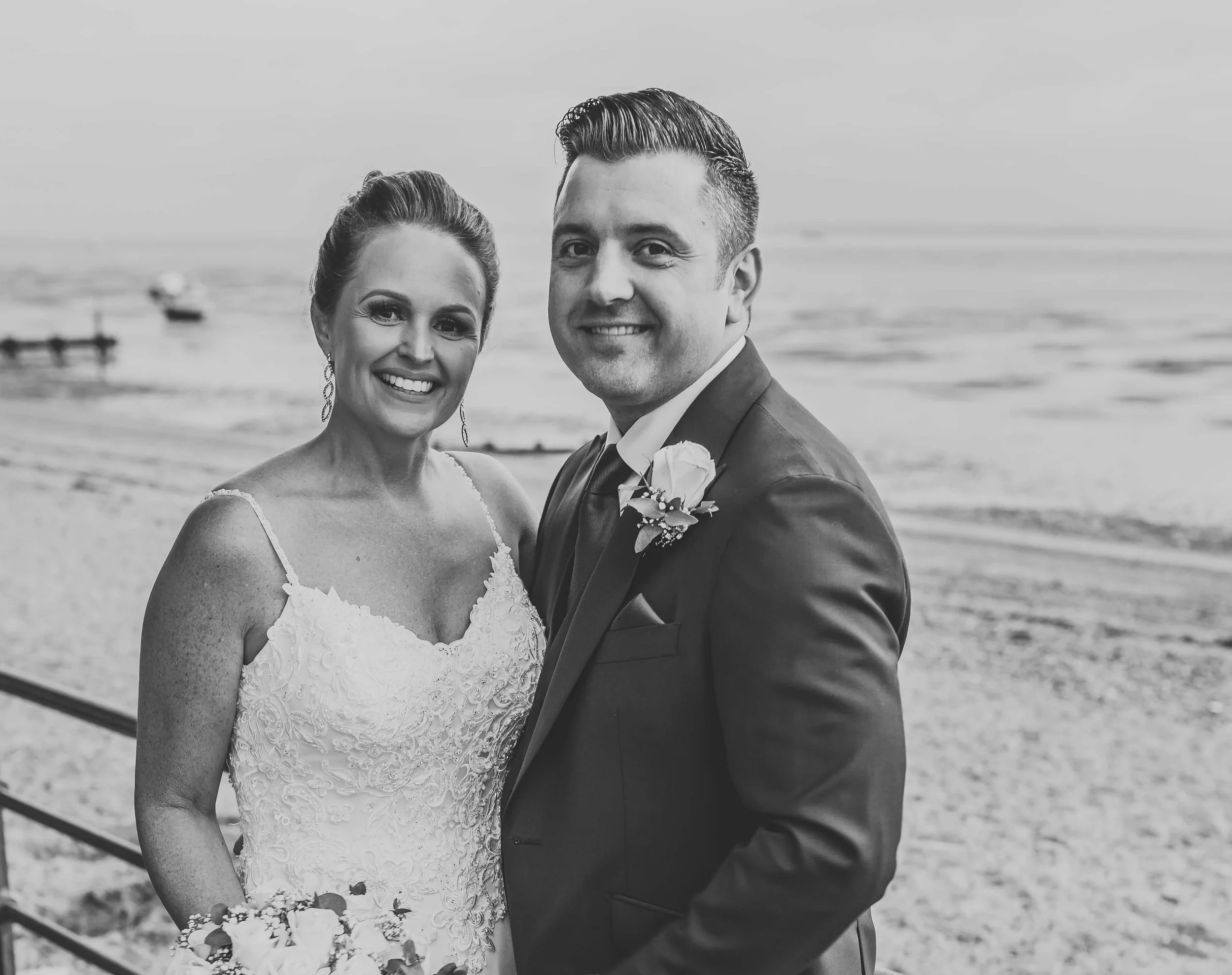 A black and white photo of a bride and groom standing on a beach, smiling.