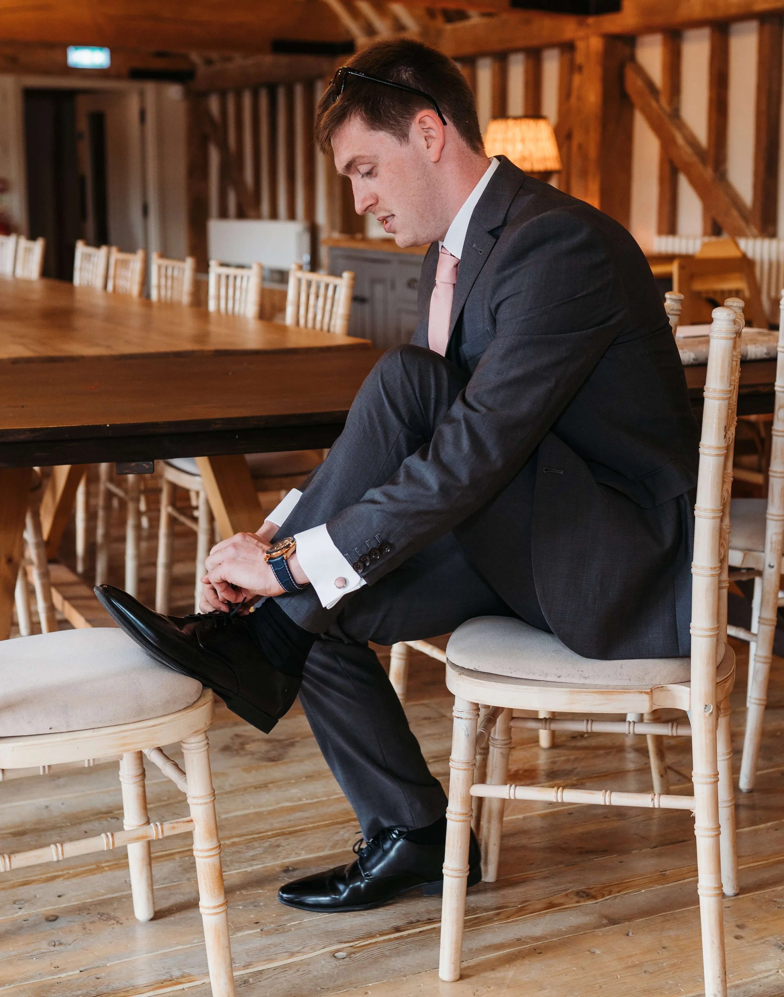 A man in a formal suit and pink tie sitting on a wooden chair, putting on black dress shoes in a rustic, wood-paneled room.