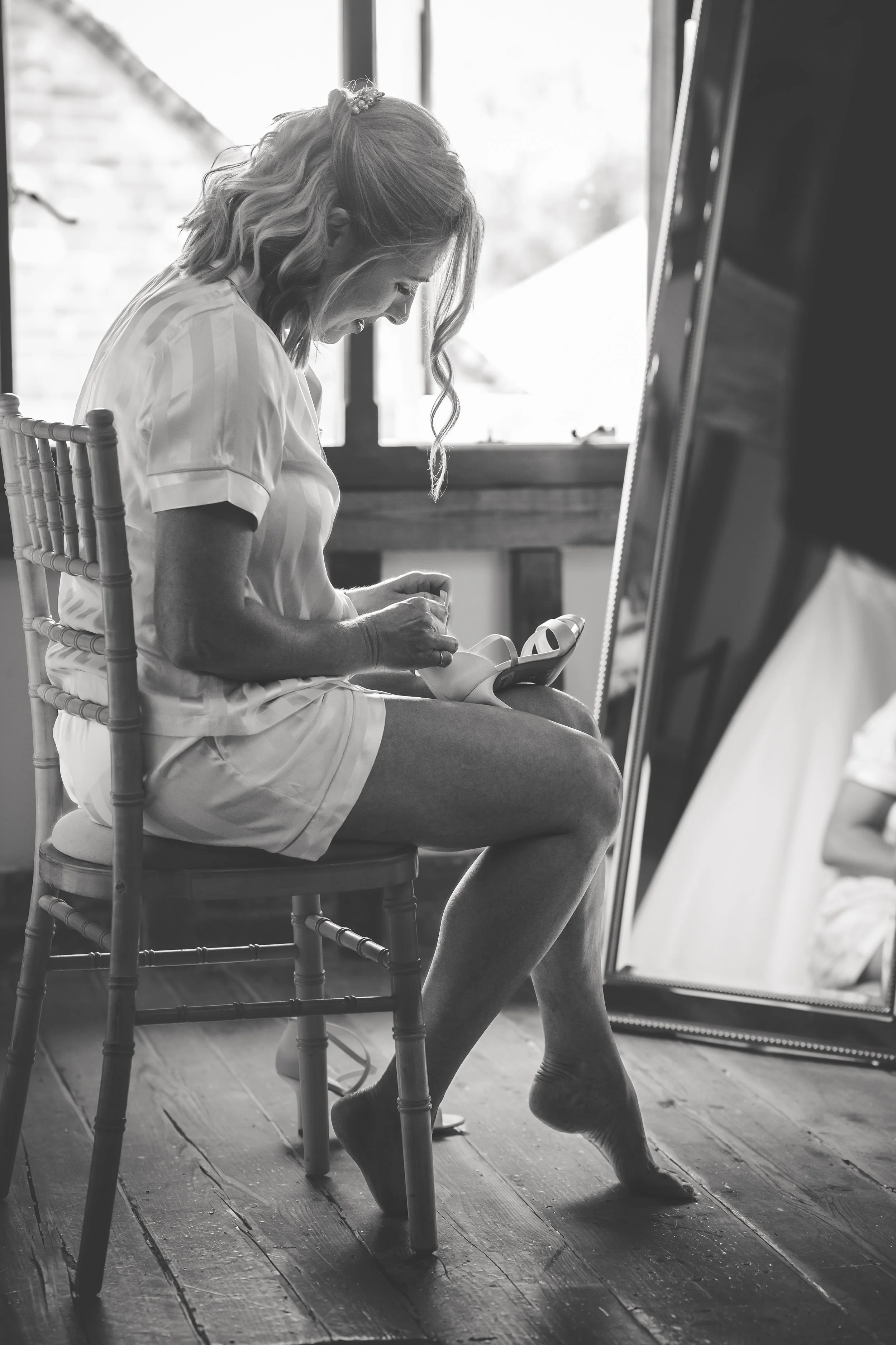 A woman in pajamas sitting on a wooden chair with her right foot elevated, holding and examining a shoe, with a full-length mirror behind her reflecting her legs and the shoe.