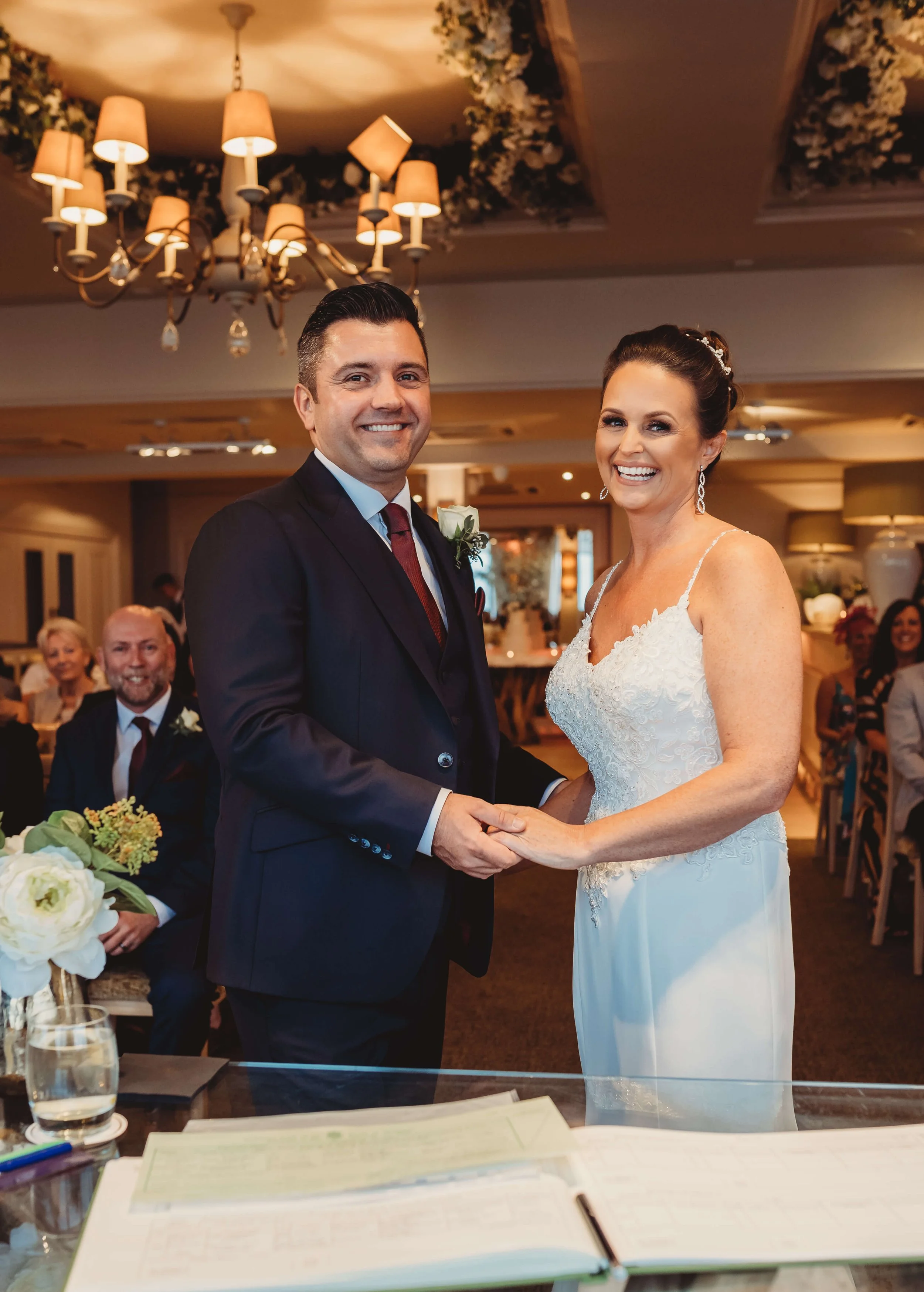 A bride and groom exchanging wedding vows indoors with smiling guests watching.