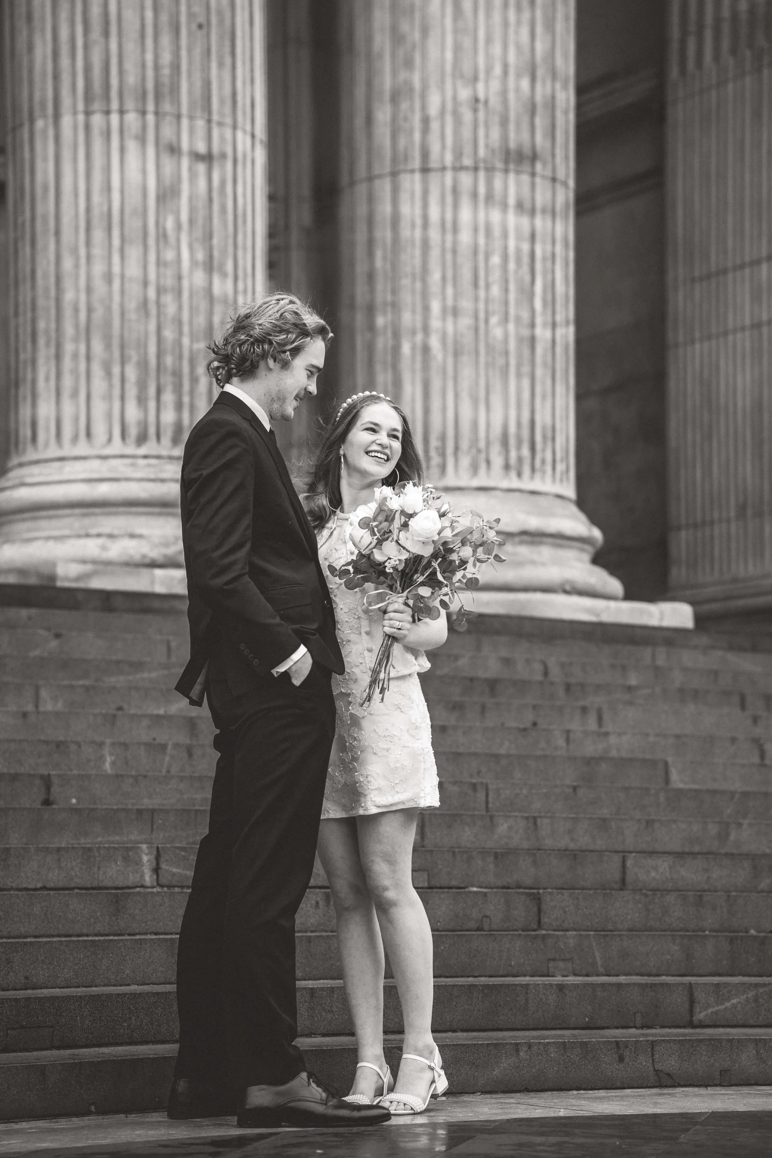 A black and white photo of a smiling bride holding a bouquet of flowers and standing on stairs next to a man in a suit, likely at a wedding or similar event.