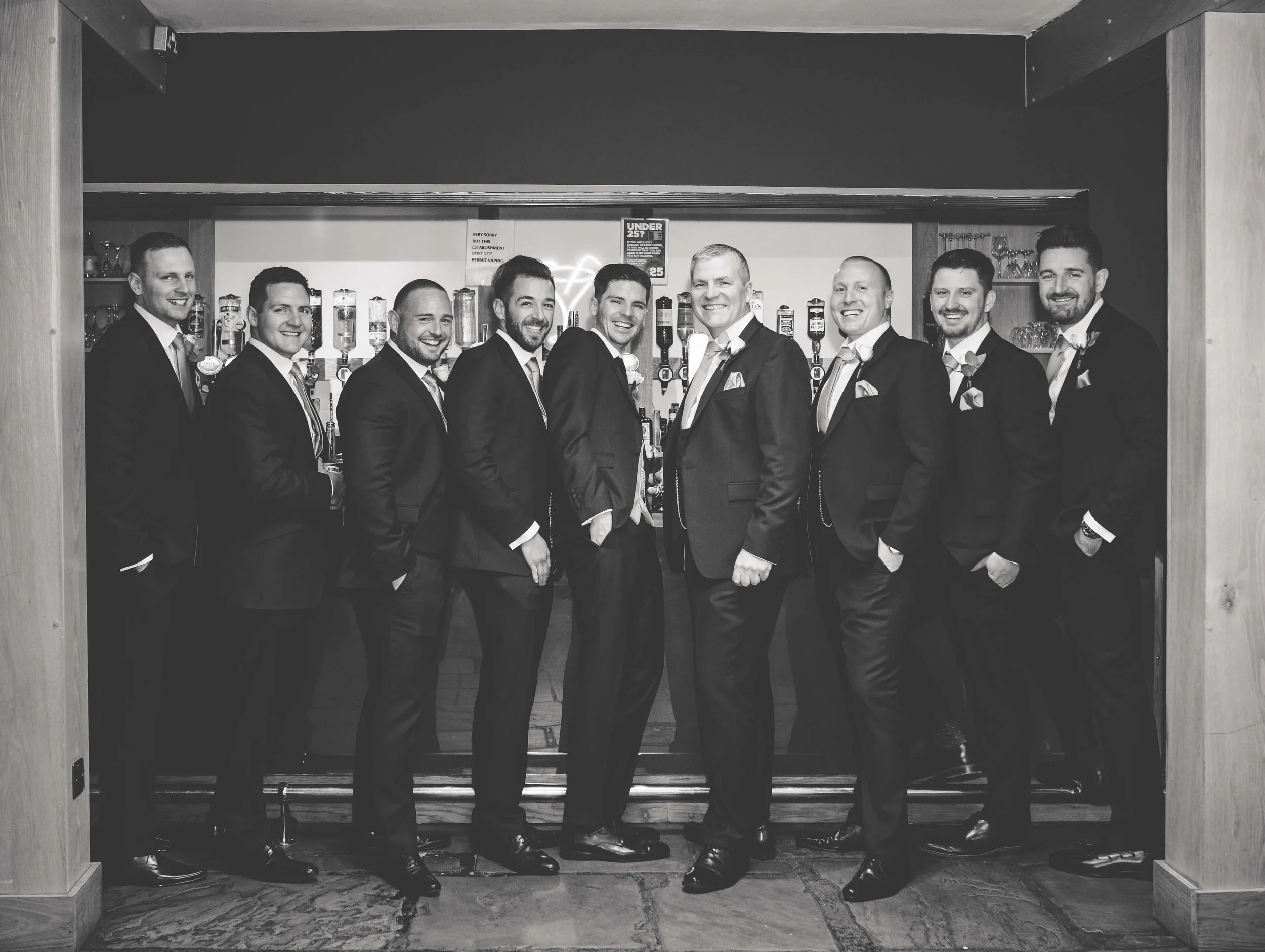 A group of ten men in matching formal suits standing together in a bar with drinks behind them, smiling for the photo.