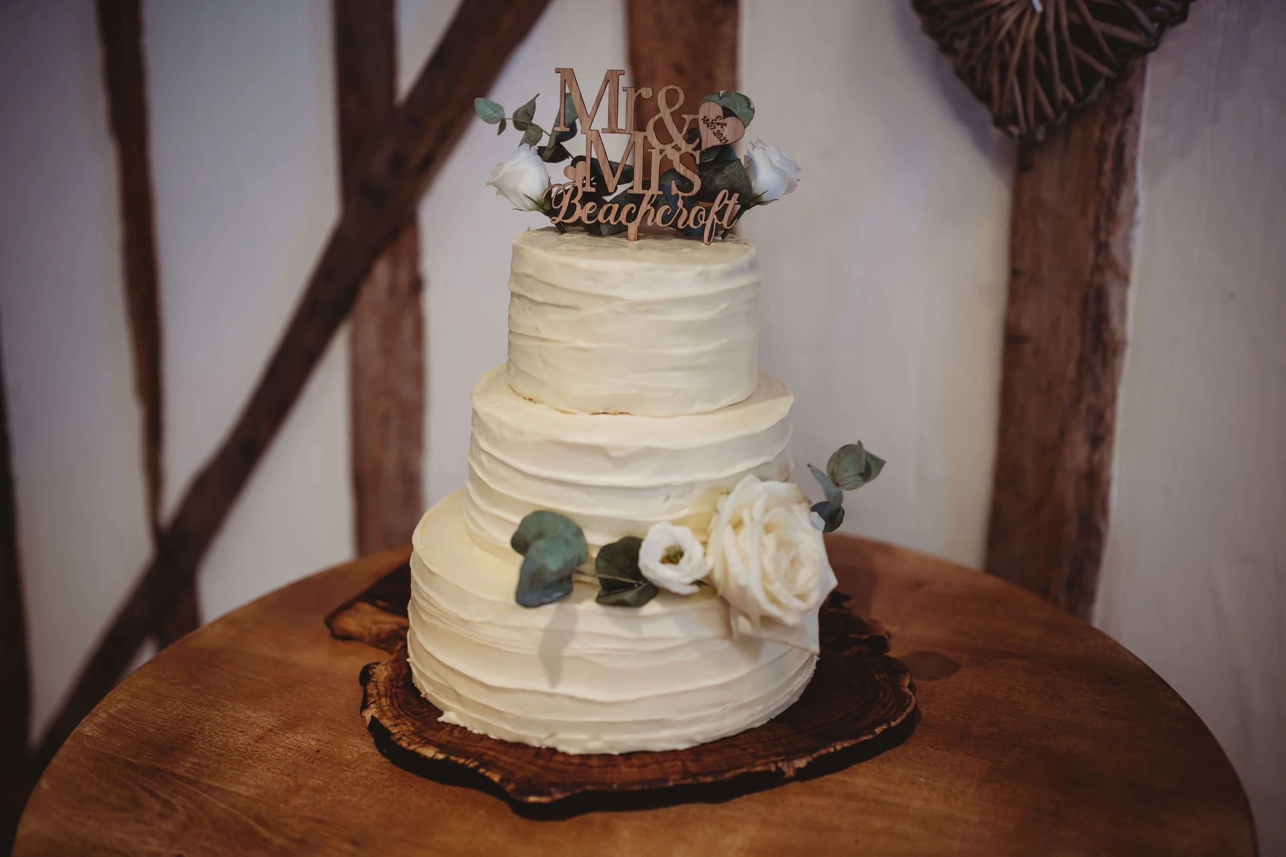 Three-tiered wedding cake with white frosting, decorated with white flowers and eucalyptus leaves, topped with a "Mrs. & Mr's Beachcroft" cake topper, set on a wooden table with a rustic wooden background.