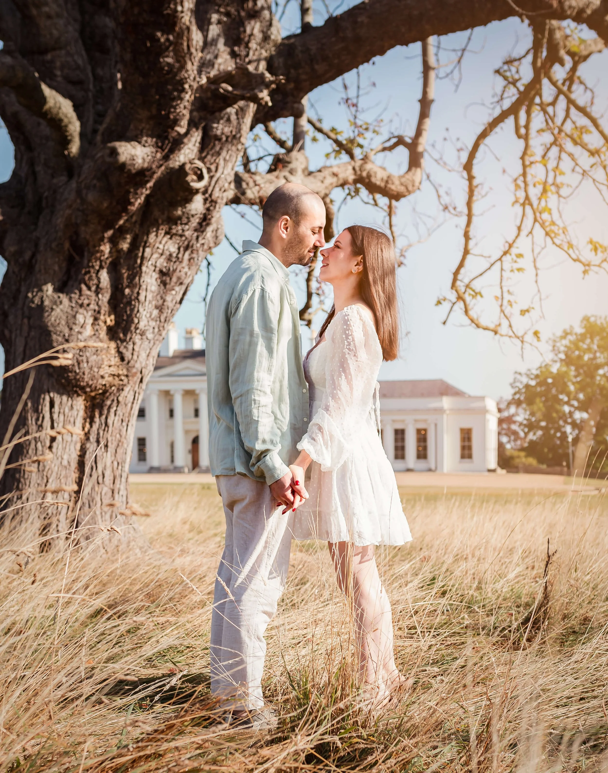 A couple holding hands and about to kiss under a large tree in a grassy field, with a white house in the background on a sunny day.