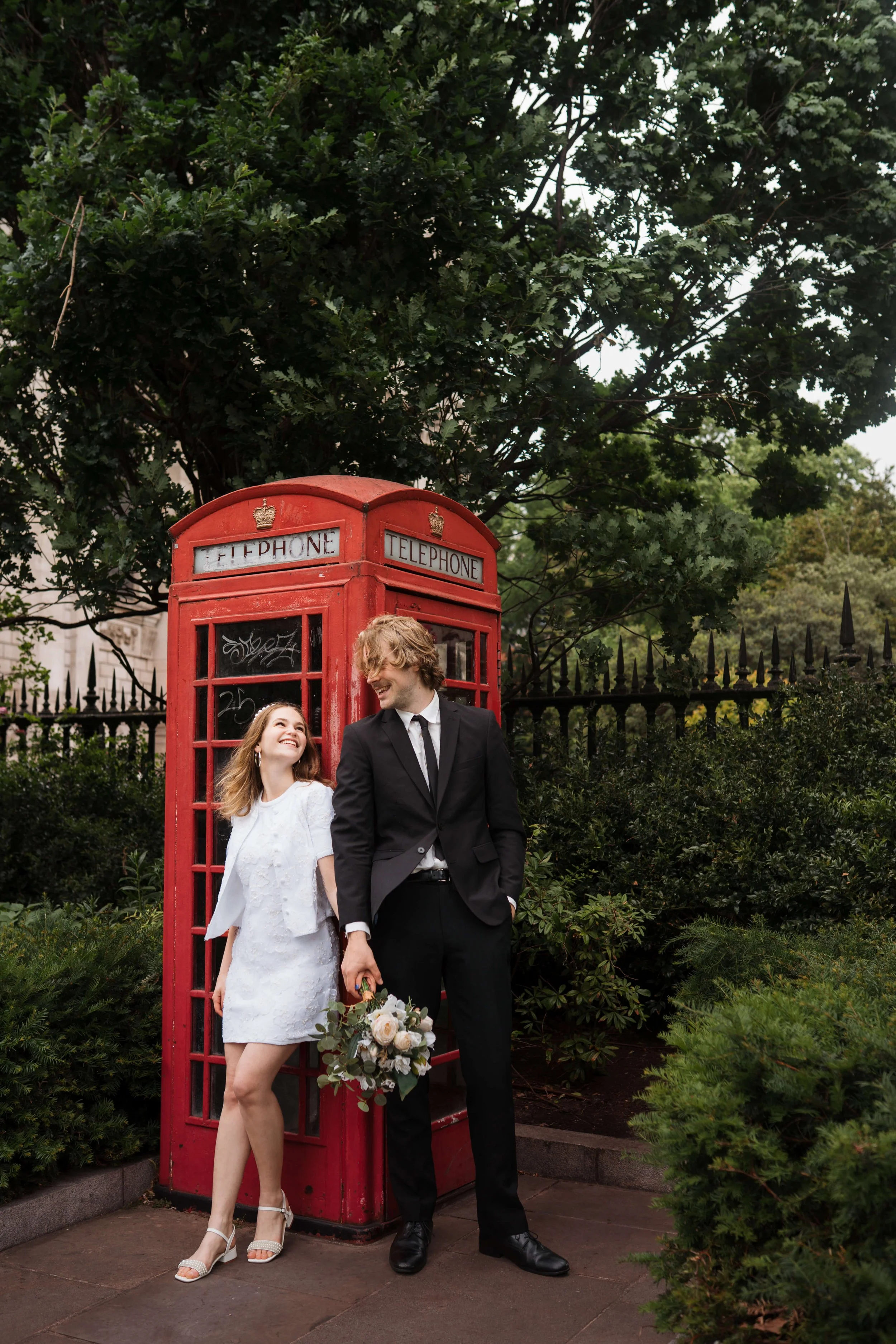 A smiling couple dressed in formal attire standing near a red British telephone booth surrounded by greenery, with the woman holding a bouquet of flowers.
