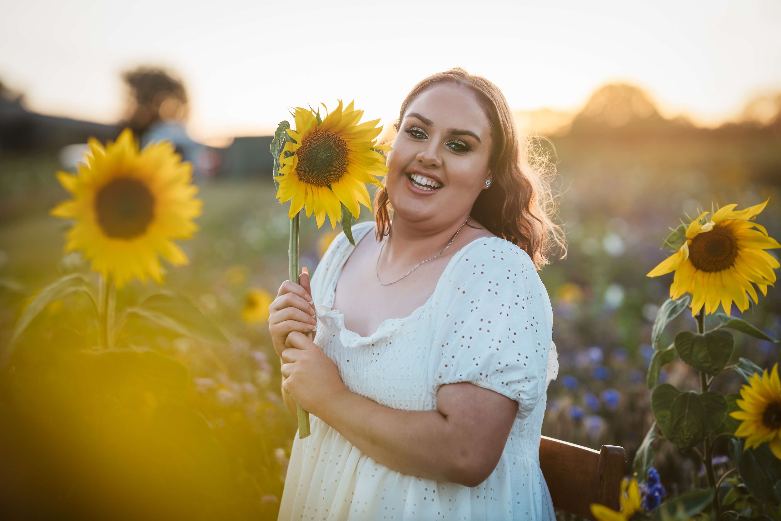 A woman in a white dress smiling and holding a sunflower in a sunflower field at sunset.