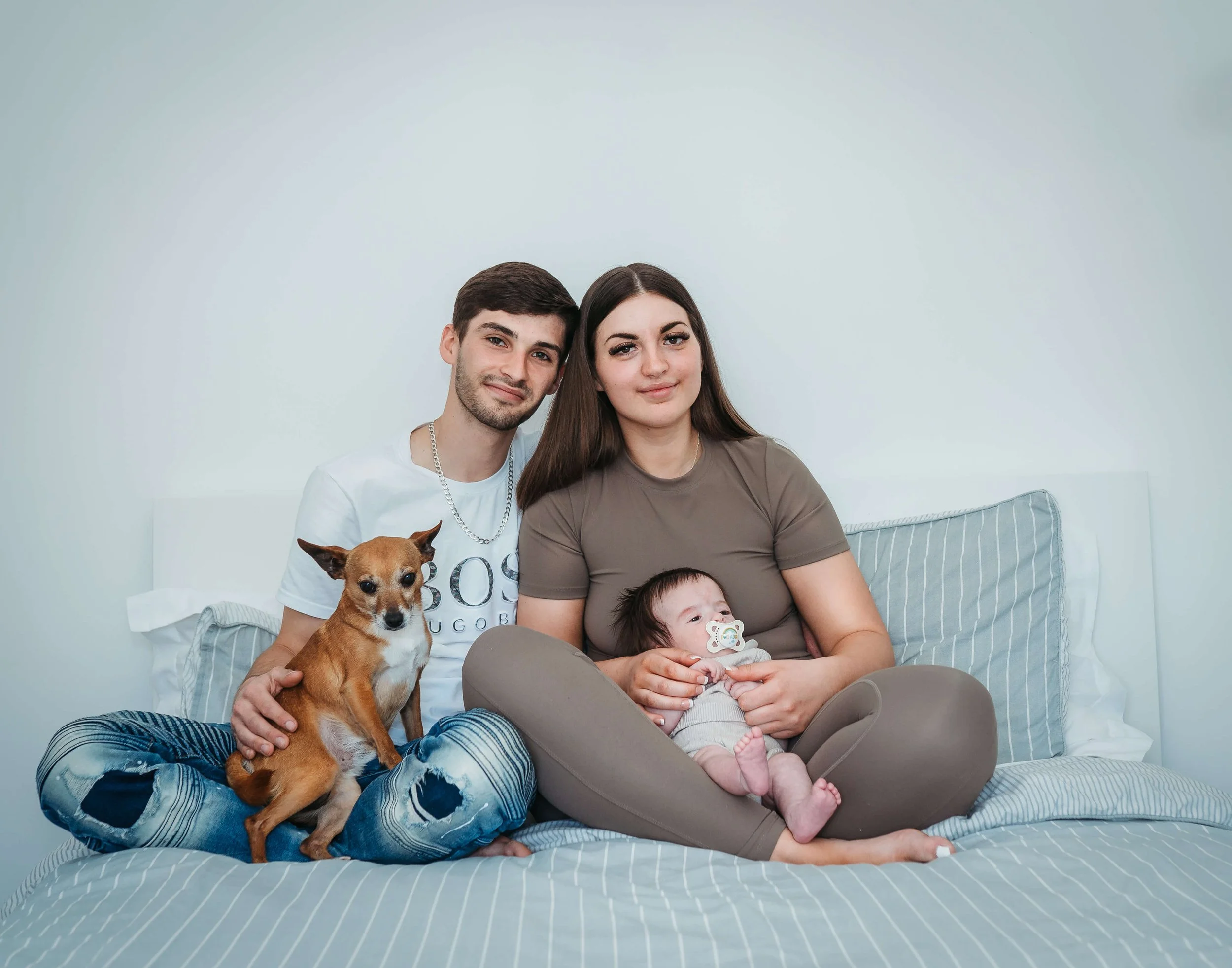 A family of four with a dog and a baby sitting on a bed in a bedroom with a white background.