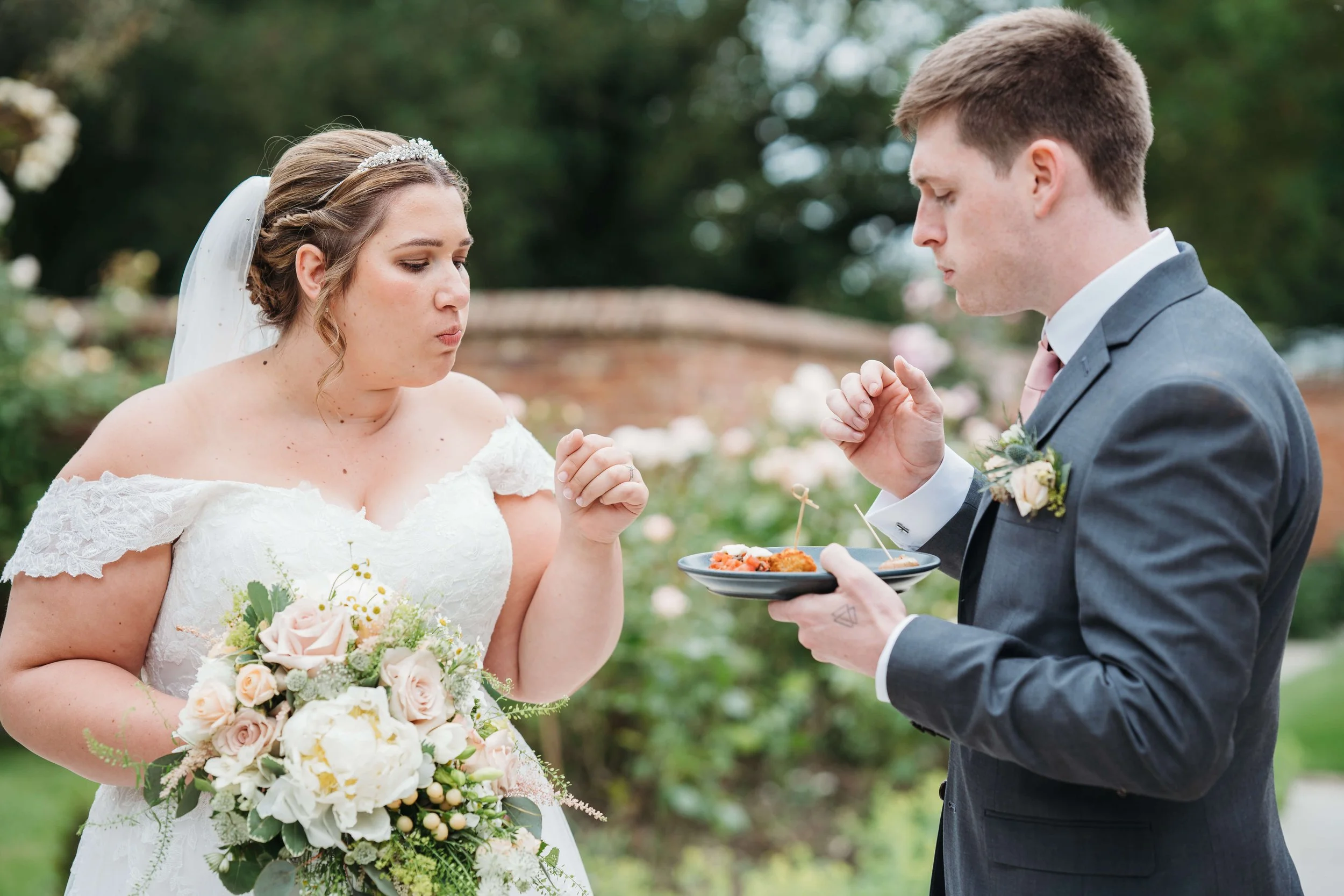 Bride in a white lace wedding dress holding a bouquet of pink and white roses and greenery, standing outside on a wedding day. Groom in a dark gray suit with boutonniere, holding a plate of food, standing across from her, with a blurred garden backgr
