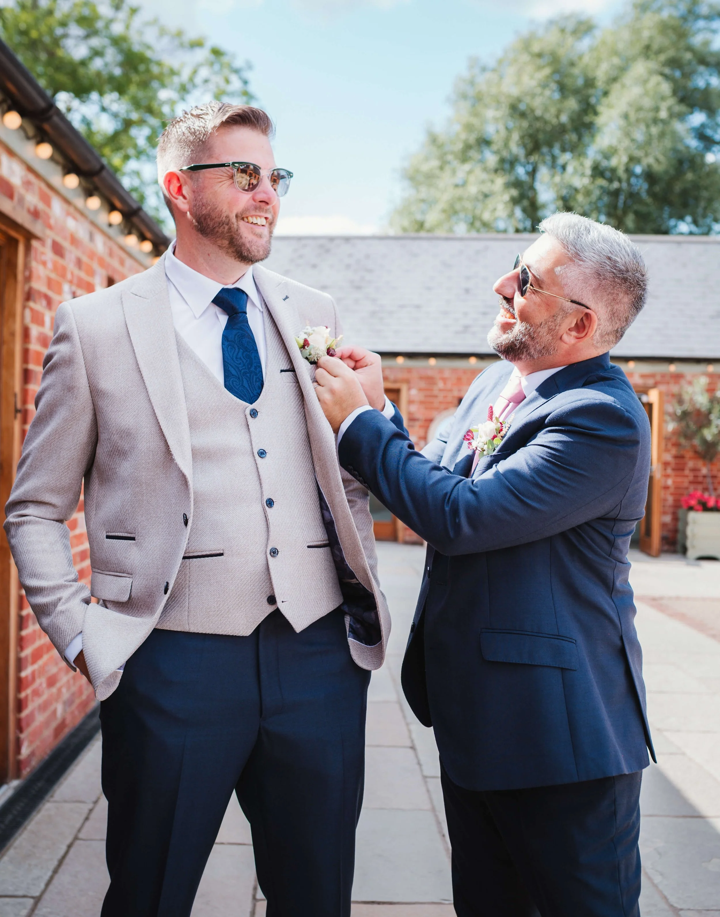 Two men in suits smiling outdoors, one adjusting the other's boutonniere during a wedding or formal event on a sunny day.