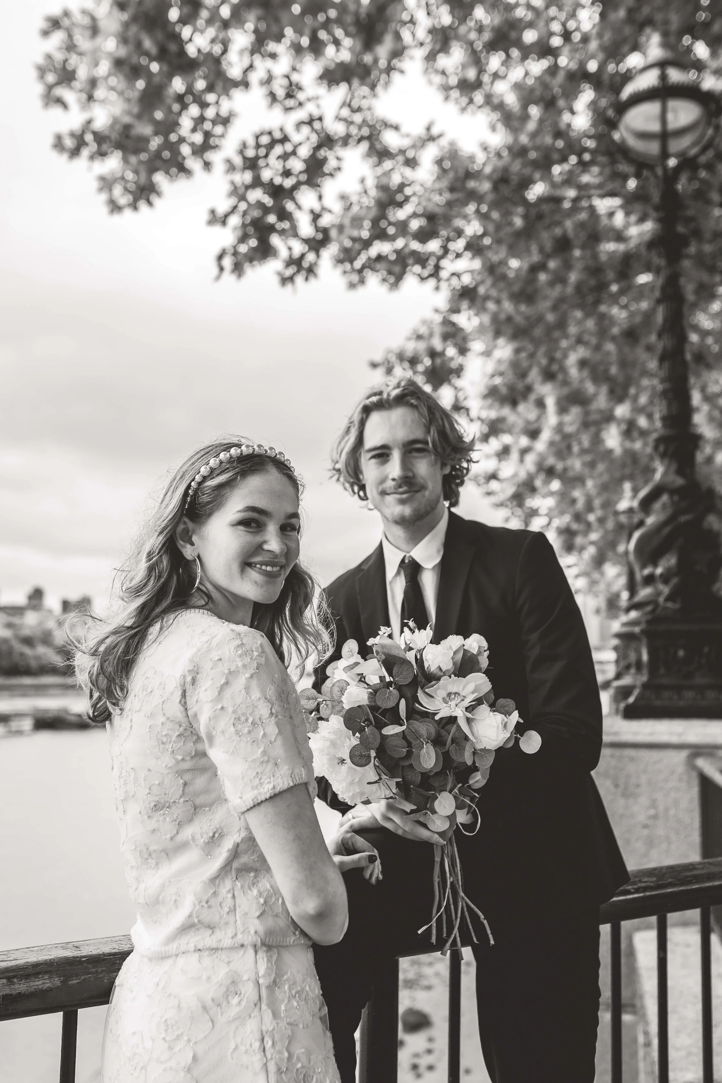 Black and white photo of a smiling woman in a lace wedding dress and a man in a suit holding a bouquet, standing outdoors near a river with trees and a lamp post in the background.