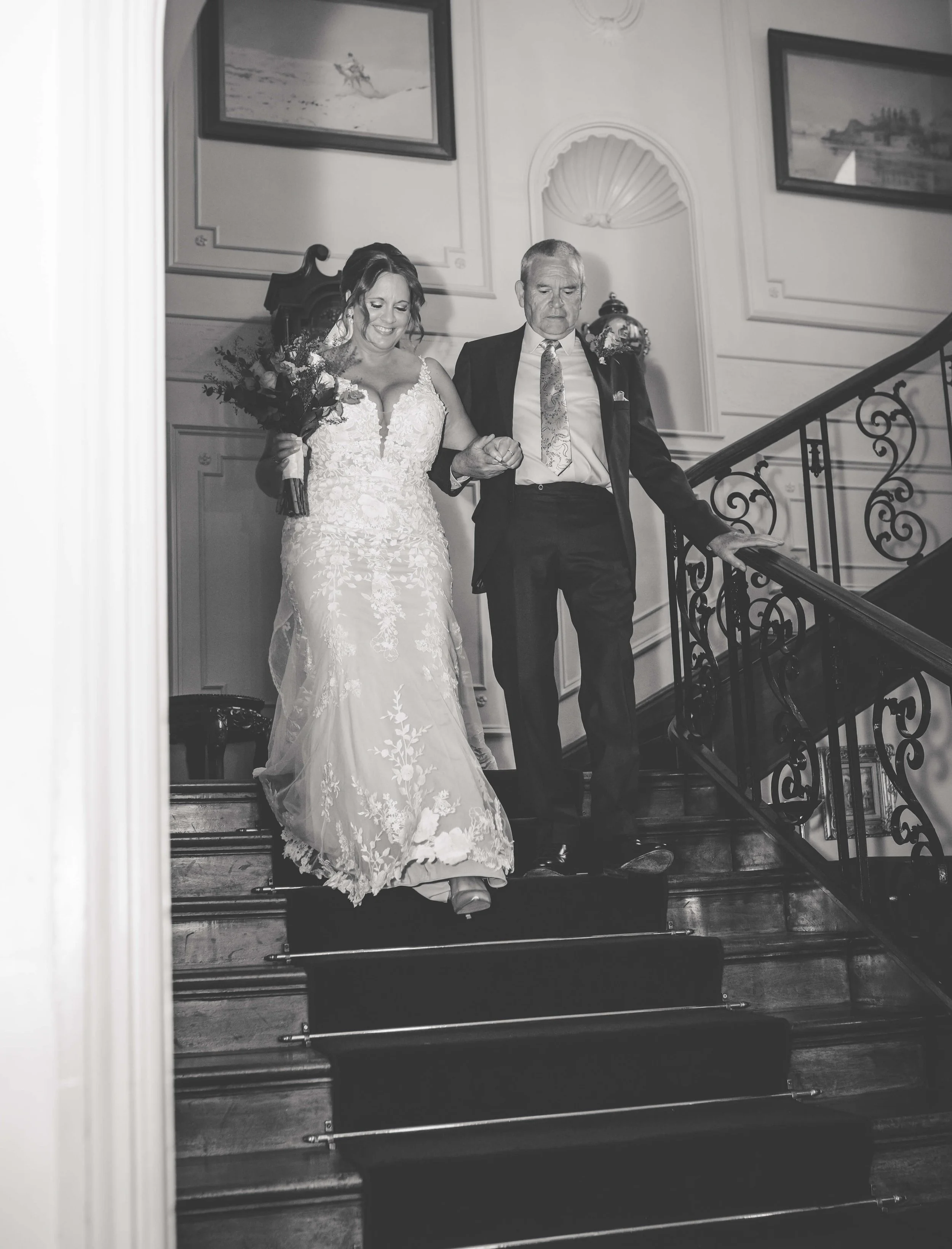 Bride in wedding dress walking down stairs with an older man, possibly her father, holding her hand, in an elegant indoor setting.