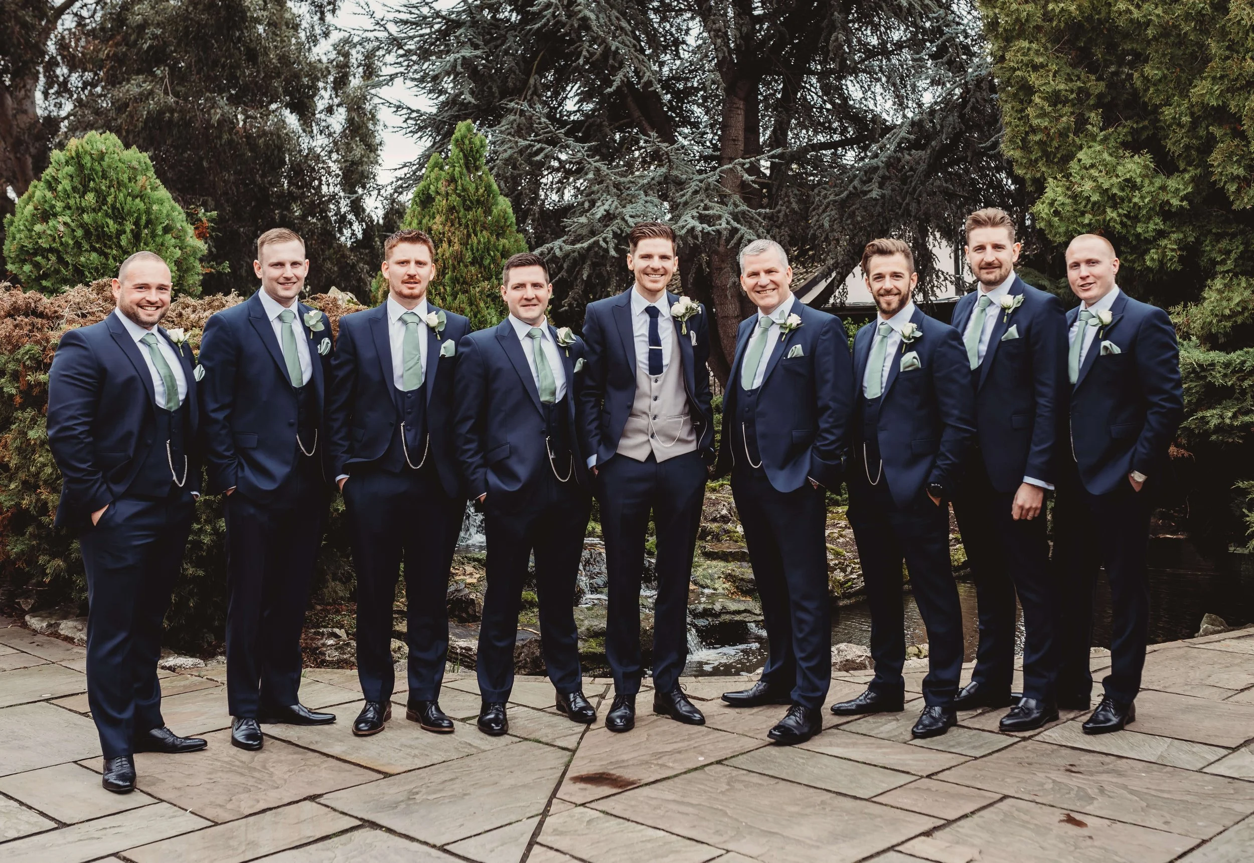 Group of groomsmen in wedding suits standing outdoors in front of trees and bushes.