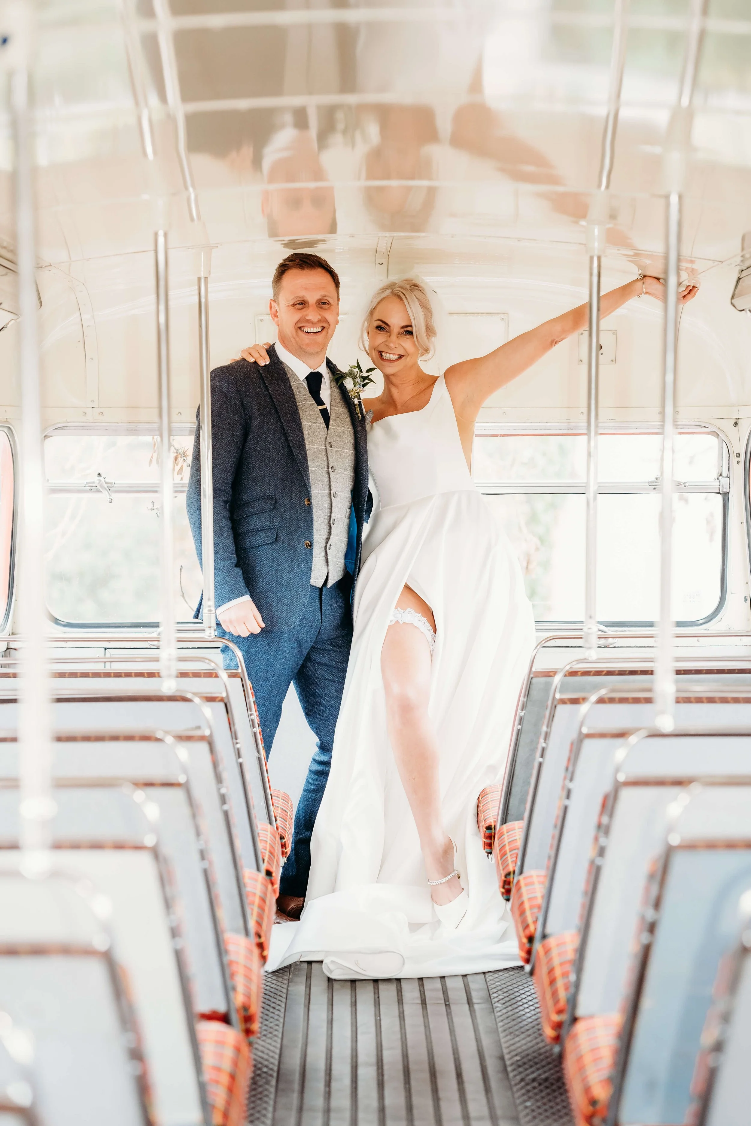 A bride and groom inside an old bus, smiling at the camera. The bride is lifting her dress, revealing her stockinged leg, and the groom has his arm around her.