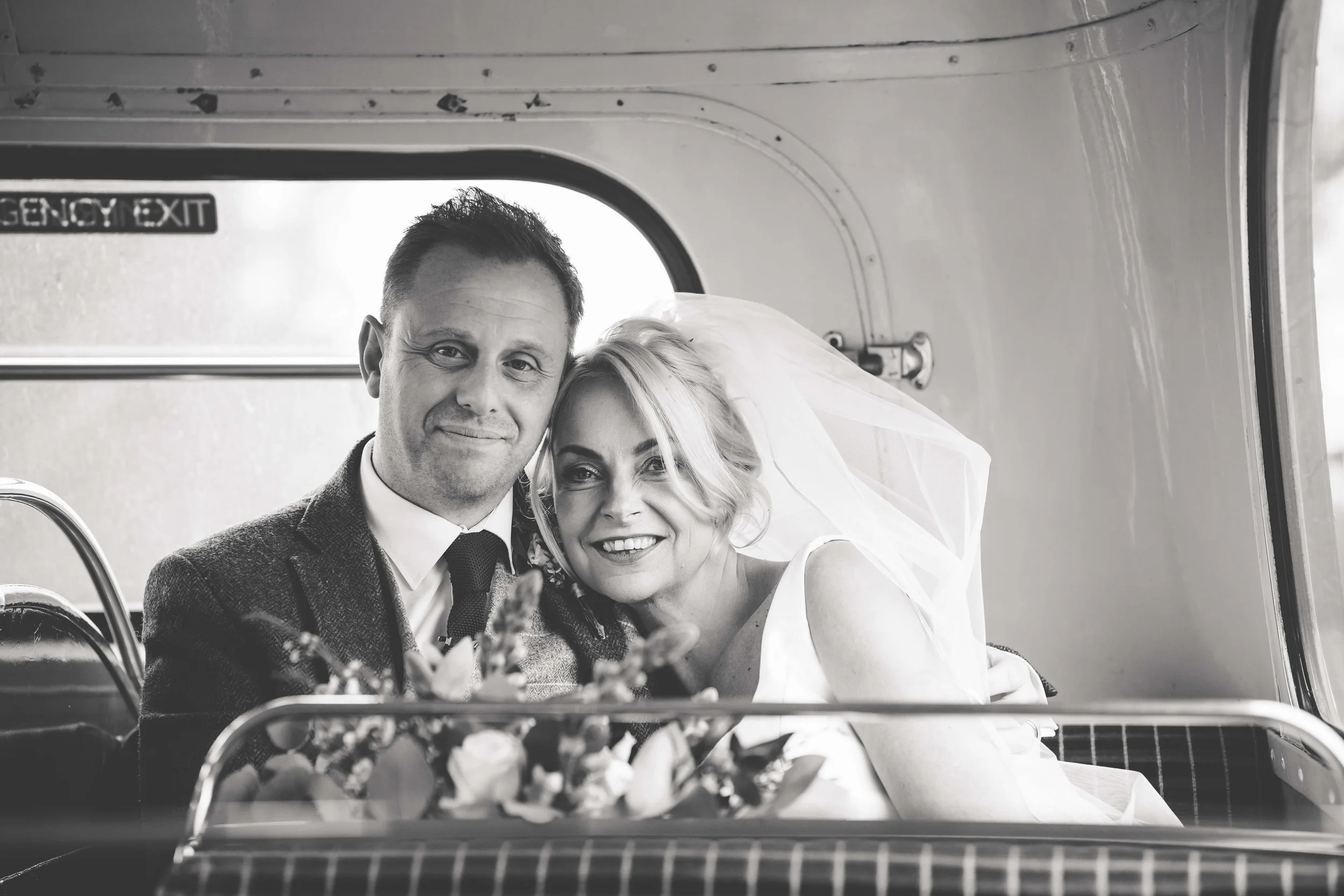 Black and white photo of a smiling man and woman dressed in wedding attire, sitting together on a bus with an 'Emergency Exit' sign visible behind them.
