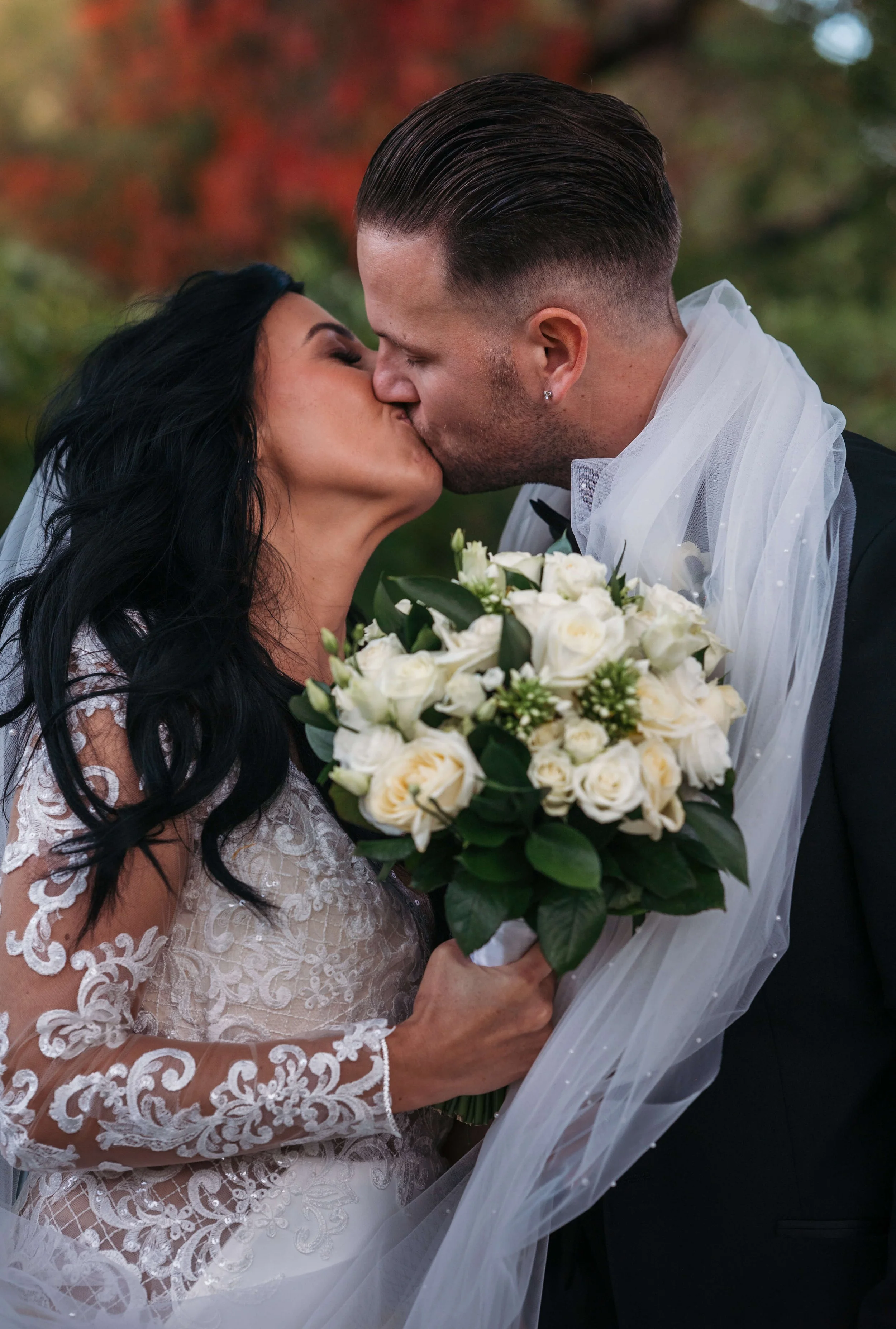 A bride and groom sharing a kiss outdoors, with the bride holding a bouquet of white roses and greenery, and the groom wearing a black suit and a white veil.
