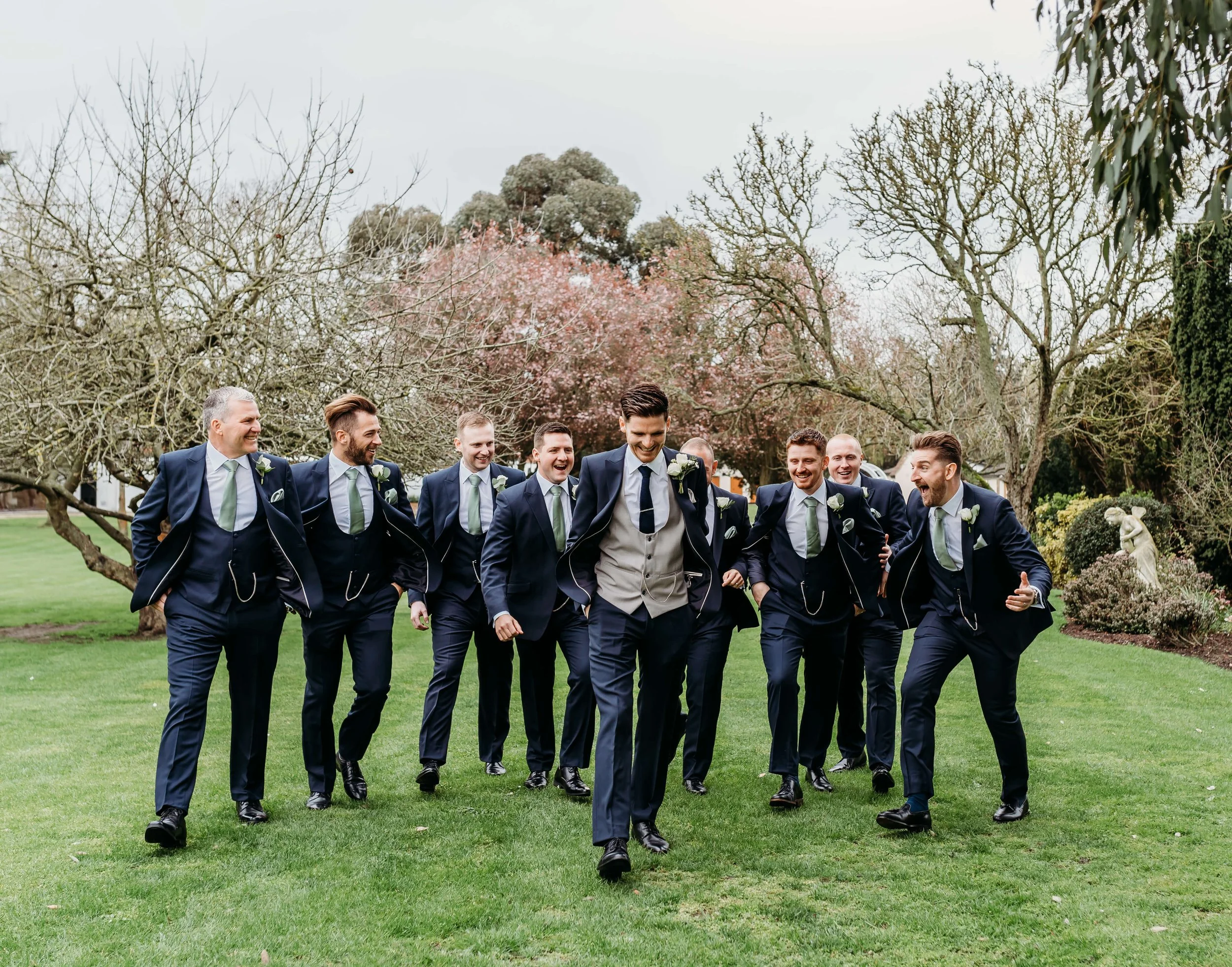 Groom and groomsmen in navy suits with light green ties, walking and laughing in a garden with trees and greenery.