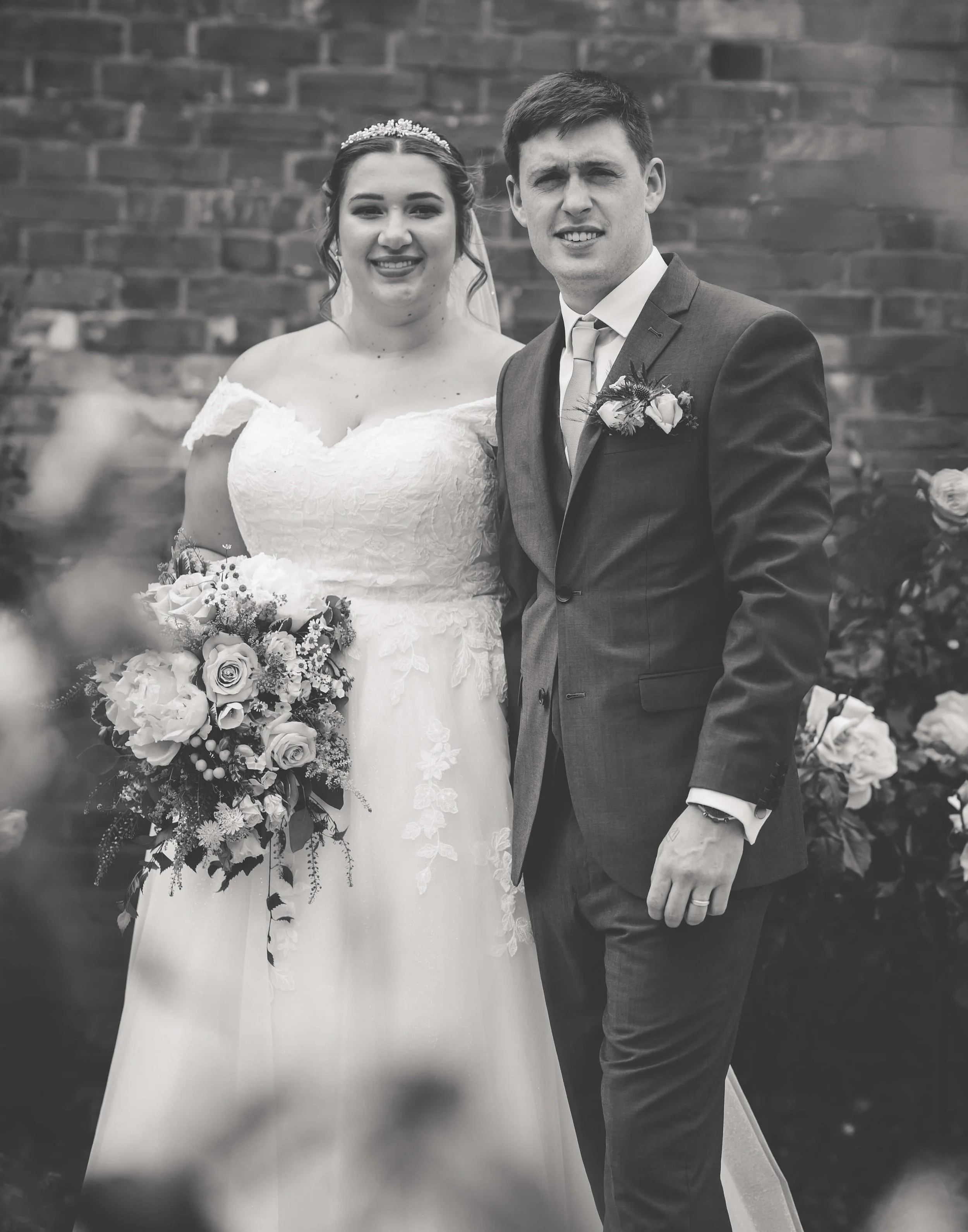 Black and white photo of a bride and groom standing together outdoors, smiling at the camera, with a brick wall in the background. The bride is holding a bouquet of flowers and wearing a lace wedding dress, while the groom is in a suit with a boutonn