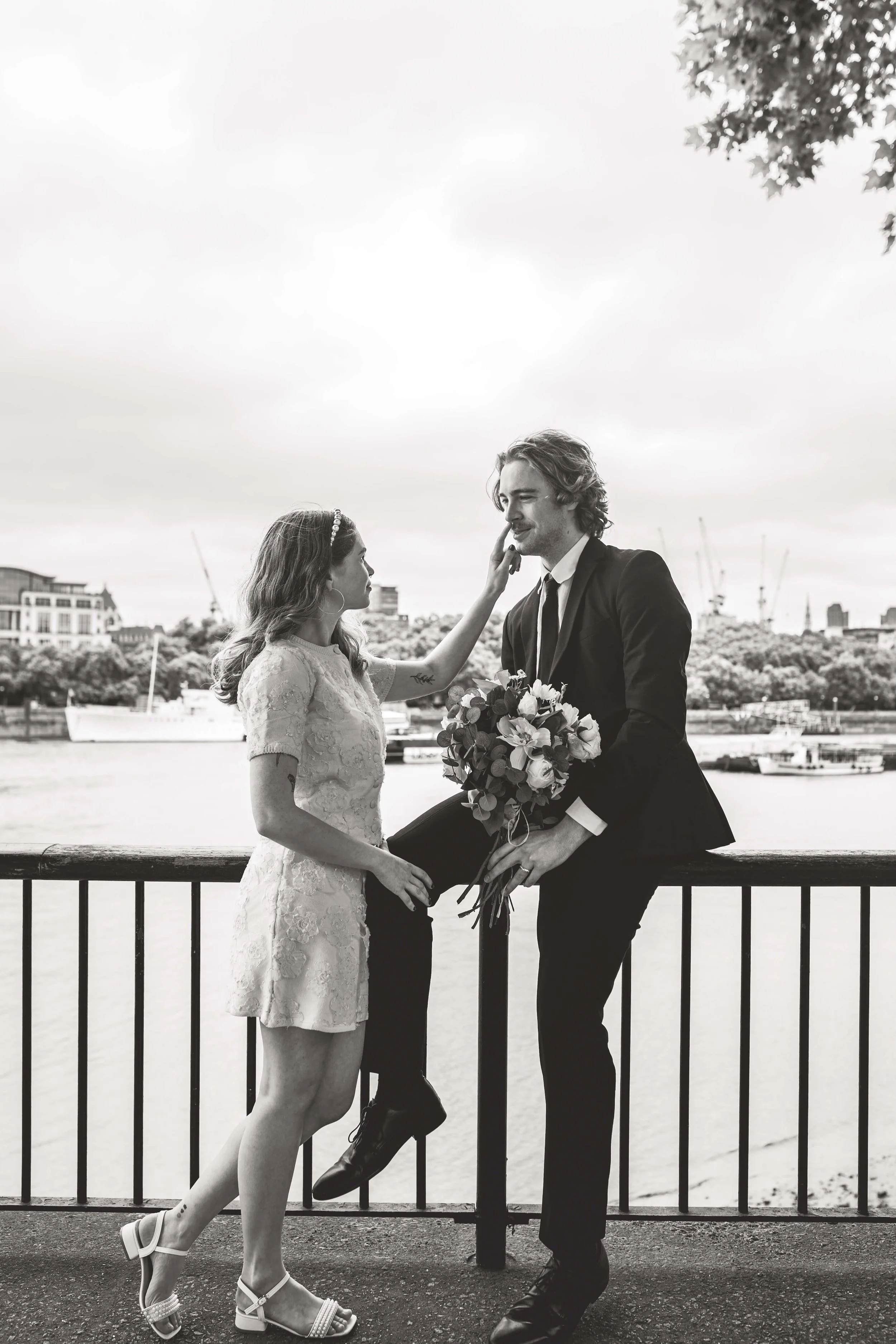 Black and white photo of a woman and a man on a bridge by a river, with boats and buildings in the background. The woman is touching the man's face and he is holding a bouquet of flowers. The woman is wearing a lace dress and heels, and the man is in