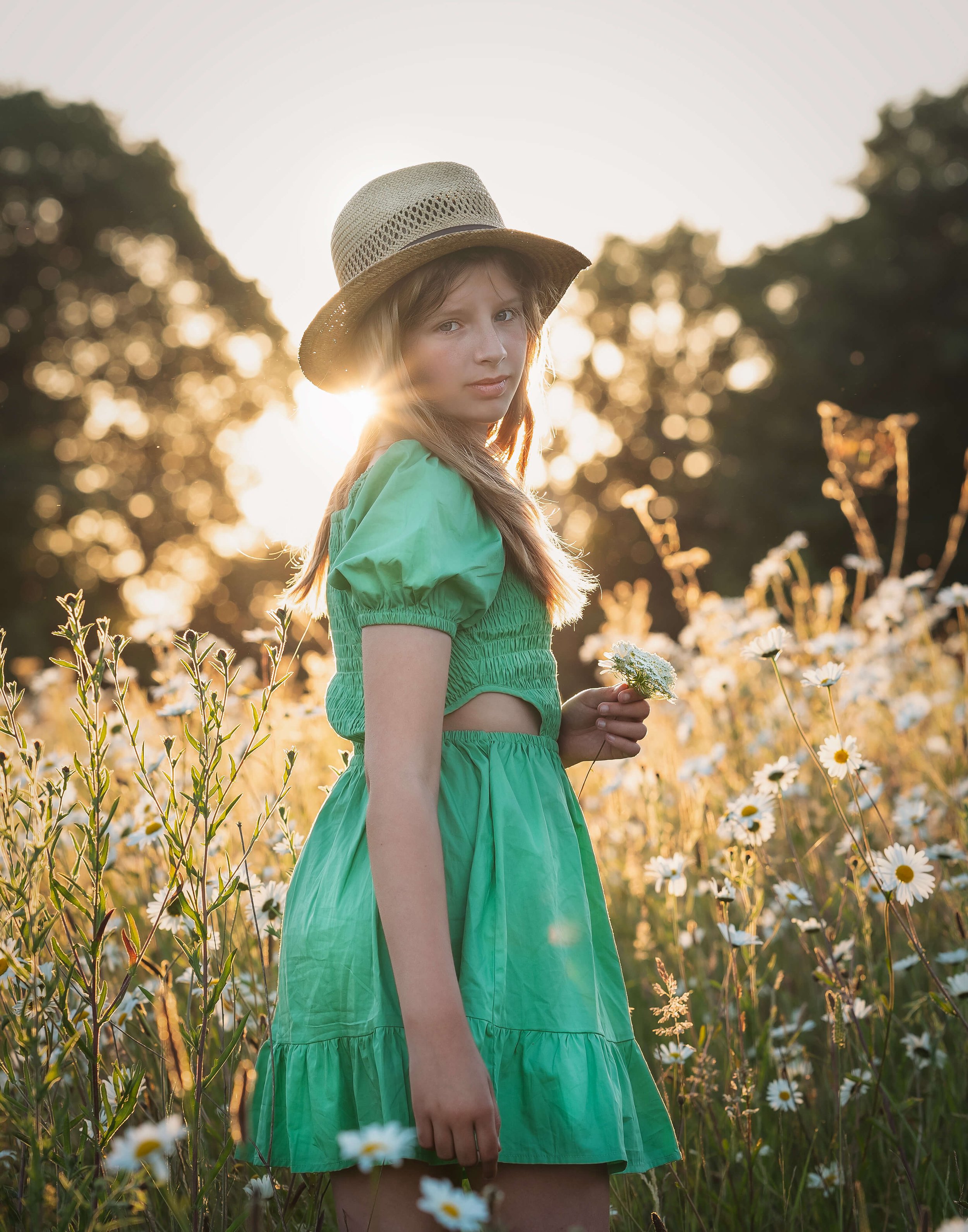 A young girl in a green dress and straw hat standing in a field of white daisies at sunset, holding a flower.