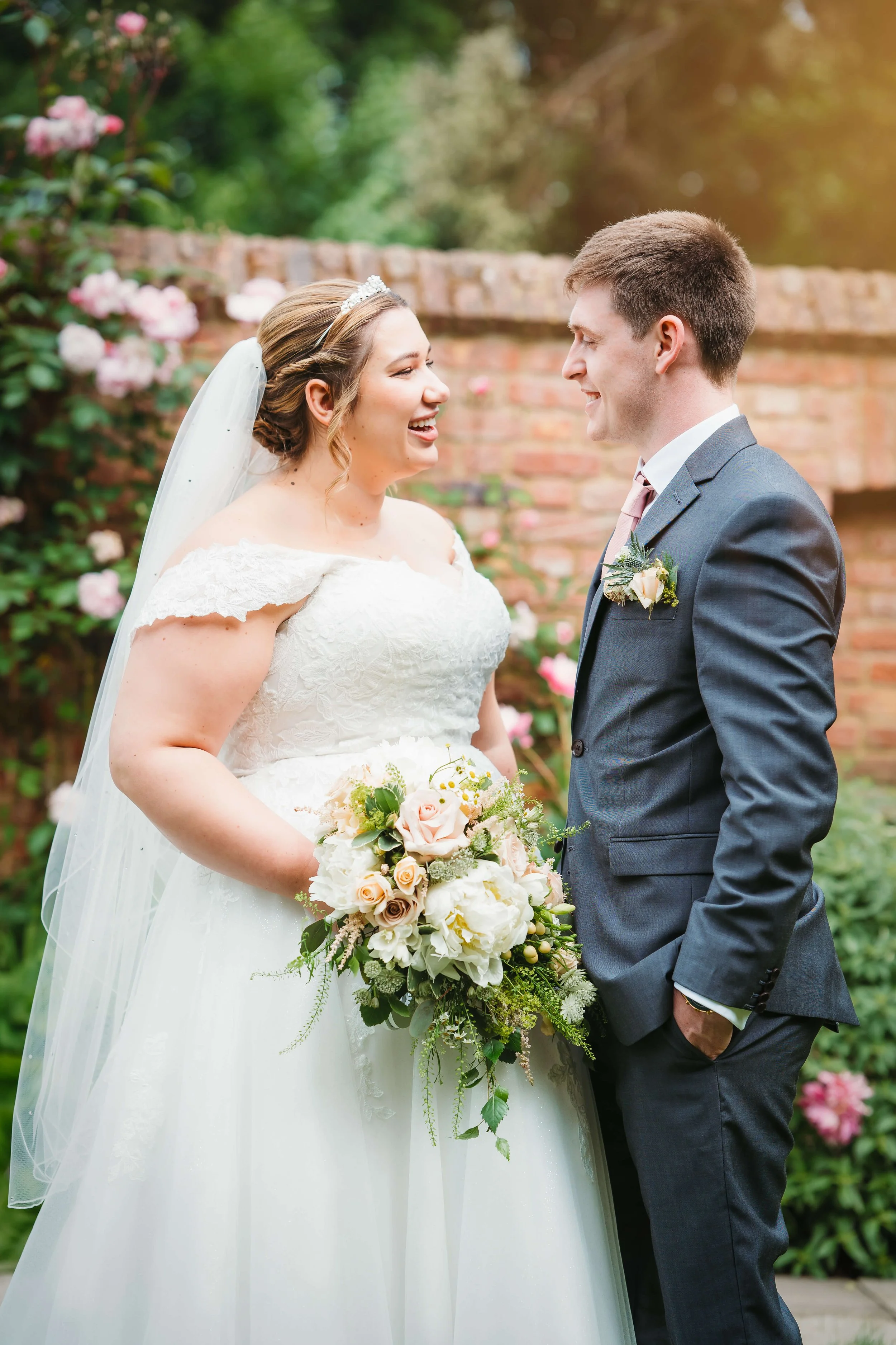 Bride and groom sharing a joyful moment outdoors, with the bride holding a bouquet, in front of a brick wall and flowers.