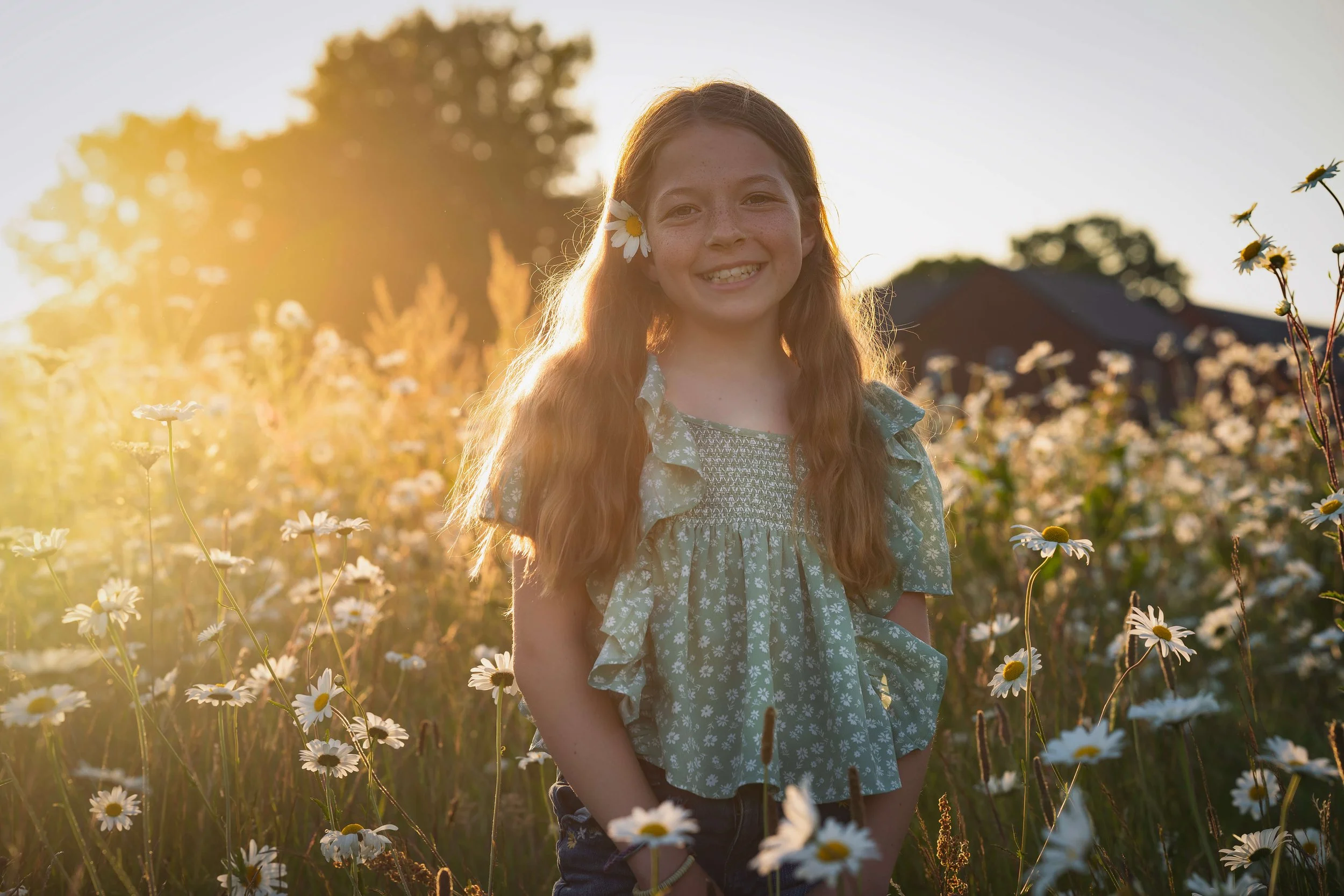 A young girl with long red hair, wearing a green floral dress, smiling in a field of daisies with the sun setting behind her.