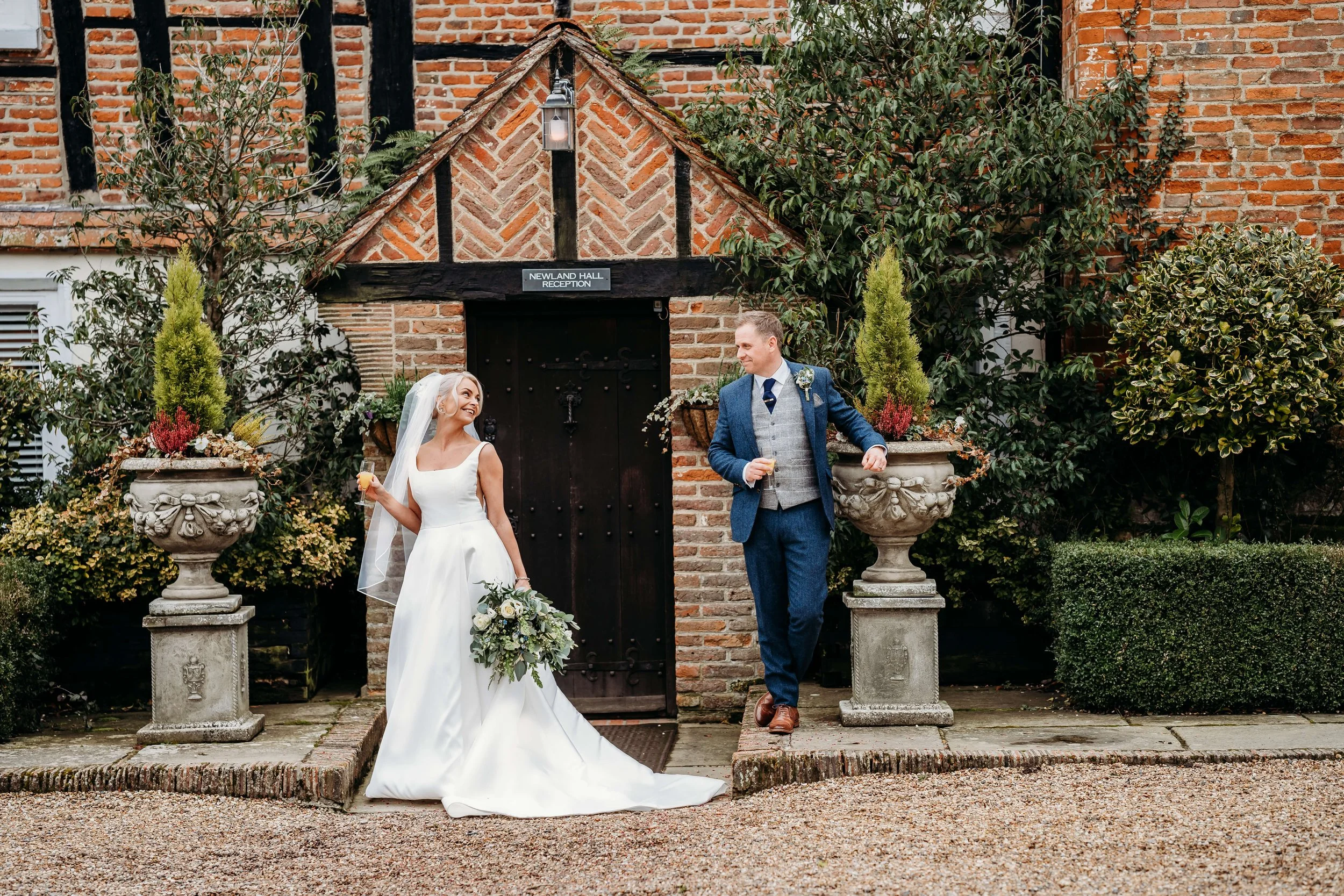 A bride and groom standing outside a brick building, smiling and holding drinks, during their wedding celebration.