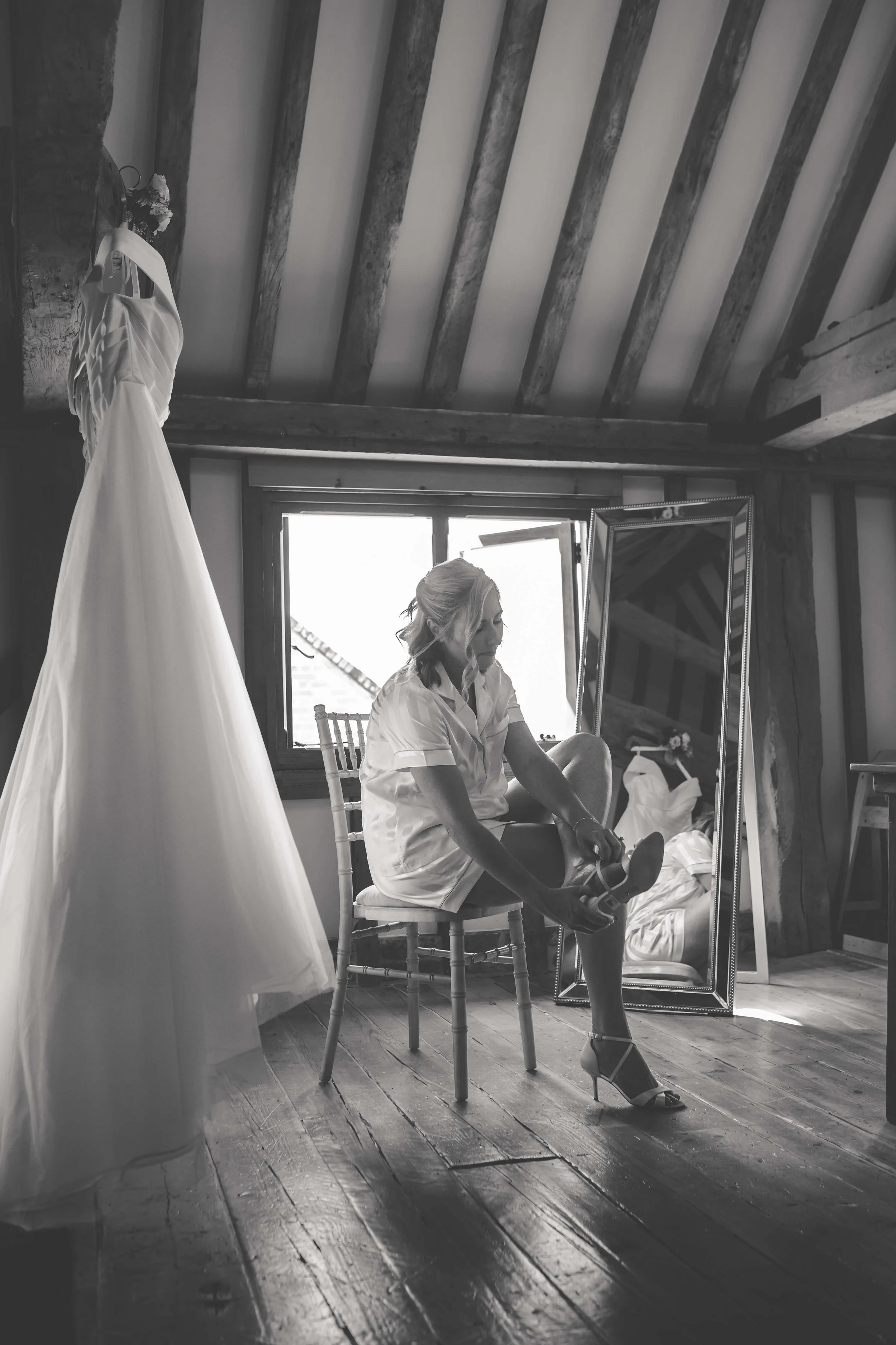A woman sitting on a chair in a rustic room, putting on high-heeled shoes, with a wedding dress hanging nearby and a full-length mirror behind her.