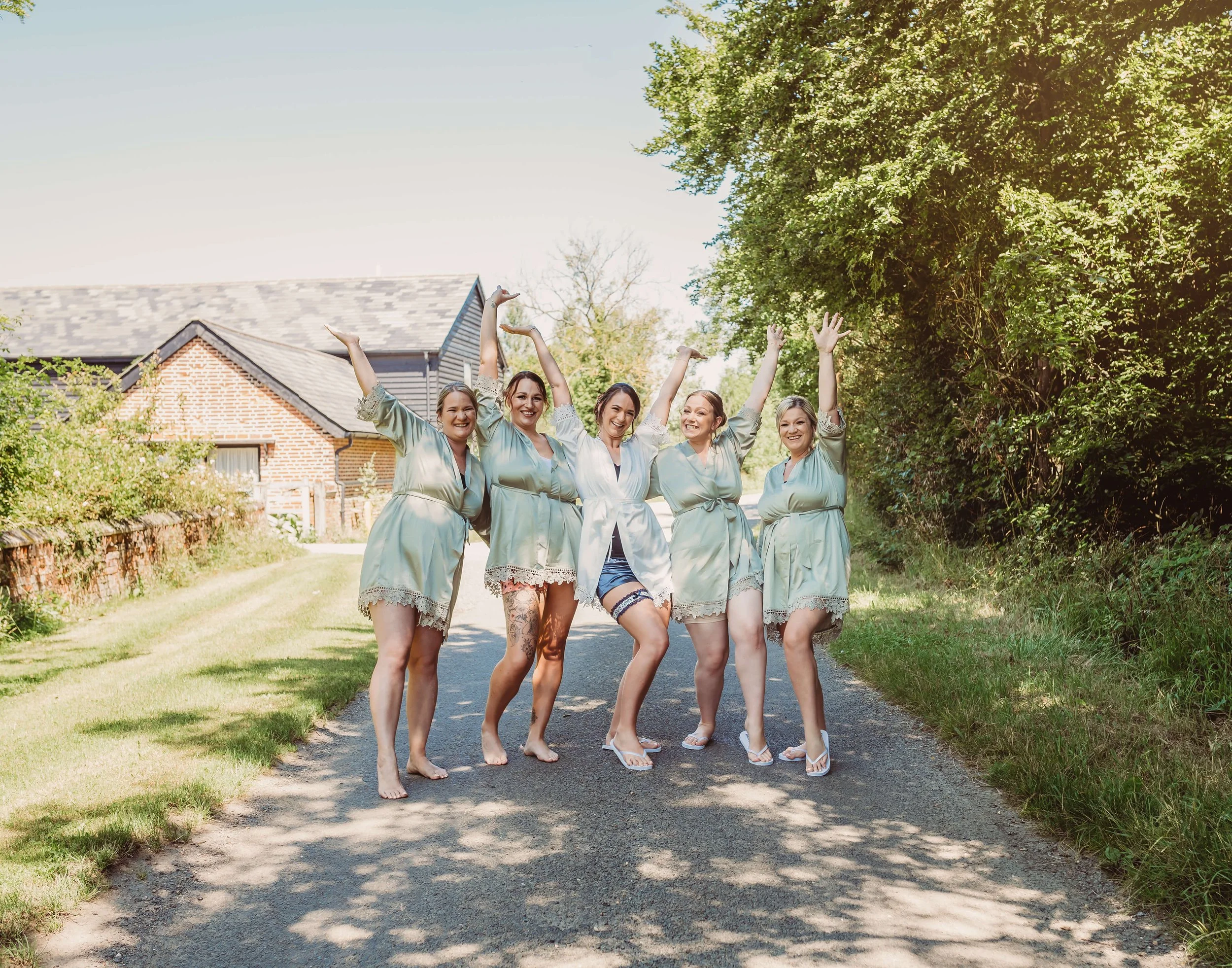 A group of five women in matching satin robes are posing barefoot on a rural road with trees and a house in the background, smiling and raising their arms in celebration.