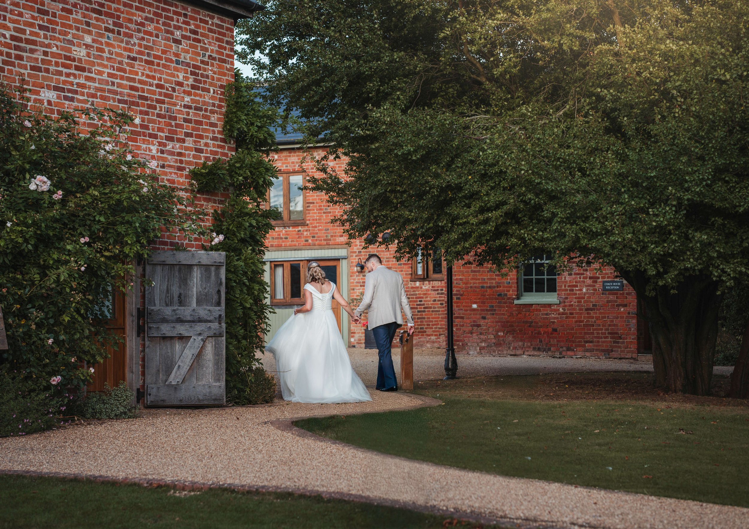 A bride and groom walking hand in hand outside a brick building, with green trees and a gravel path in the foreground.