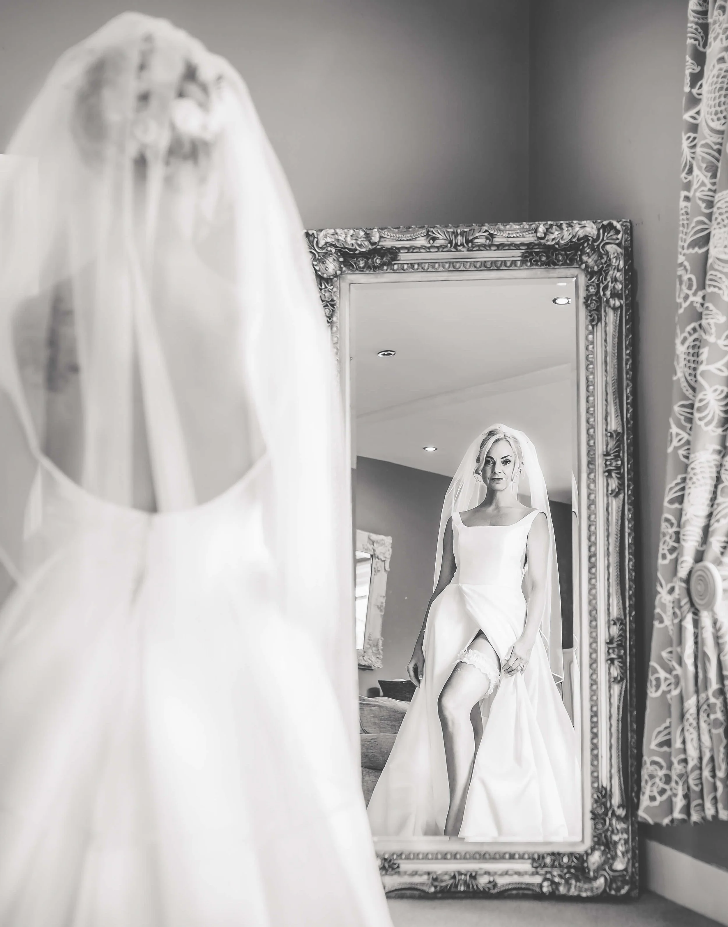 A bride wearing a wedding dress and veil, looking at herself in a large ornate mirror.
