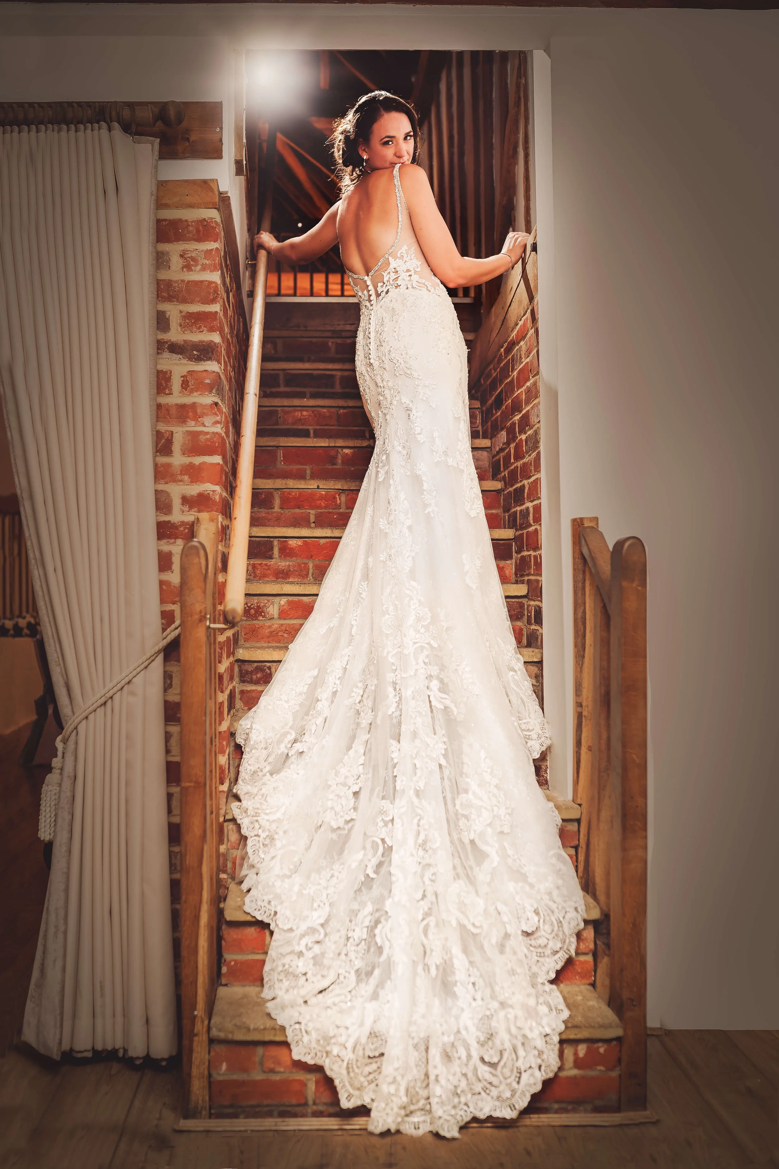 A bride in a white lace wedding gown standing on a brick staircase, looking back over her shoulder in a rustic interior setting.