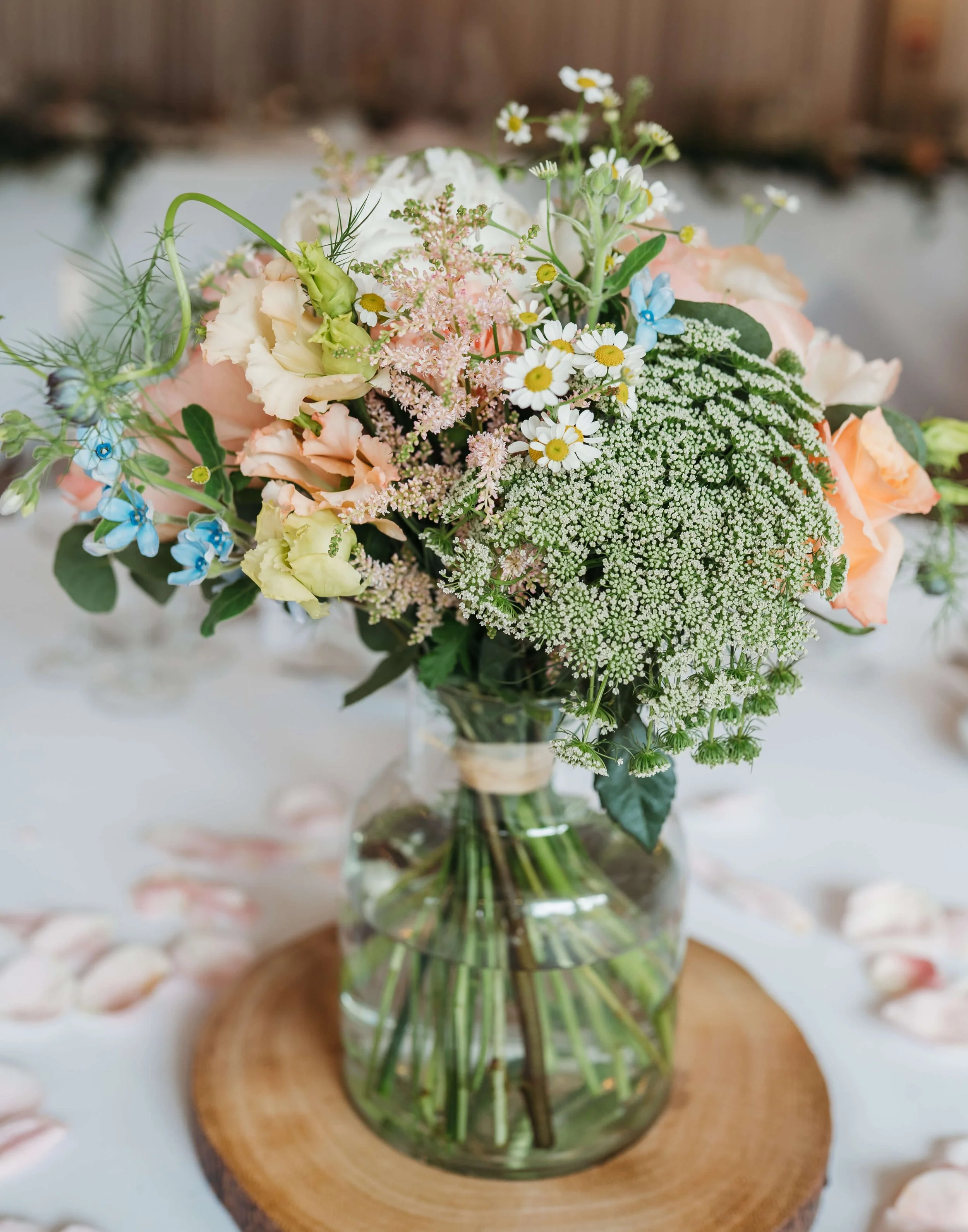 A bouquet of mixed flowers in a glass jar on a wooden base, featuring white, pink, and blue flowers with green foliage.