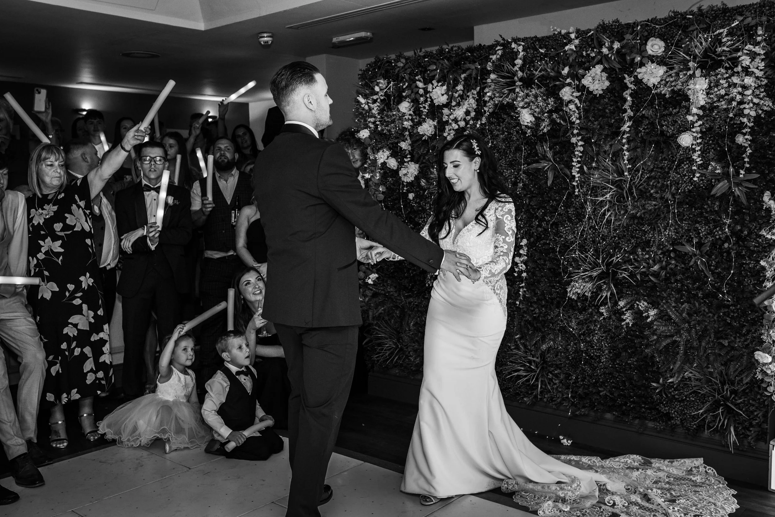 A bride and groom dance at their wedding reception, surrounded by guests who are holding candles and watching happily. The bride wears a long, elegant lace wedding gown, and the groom is in a dark suit. The background features a floral wall.