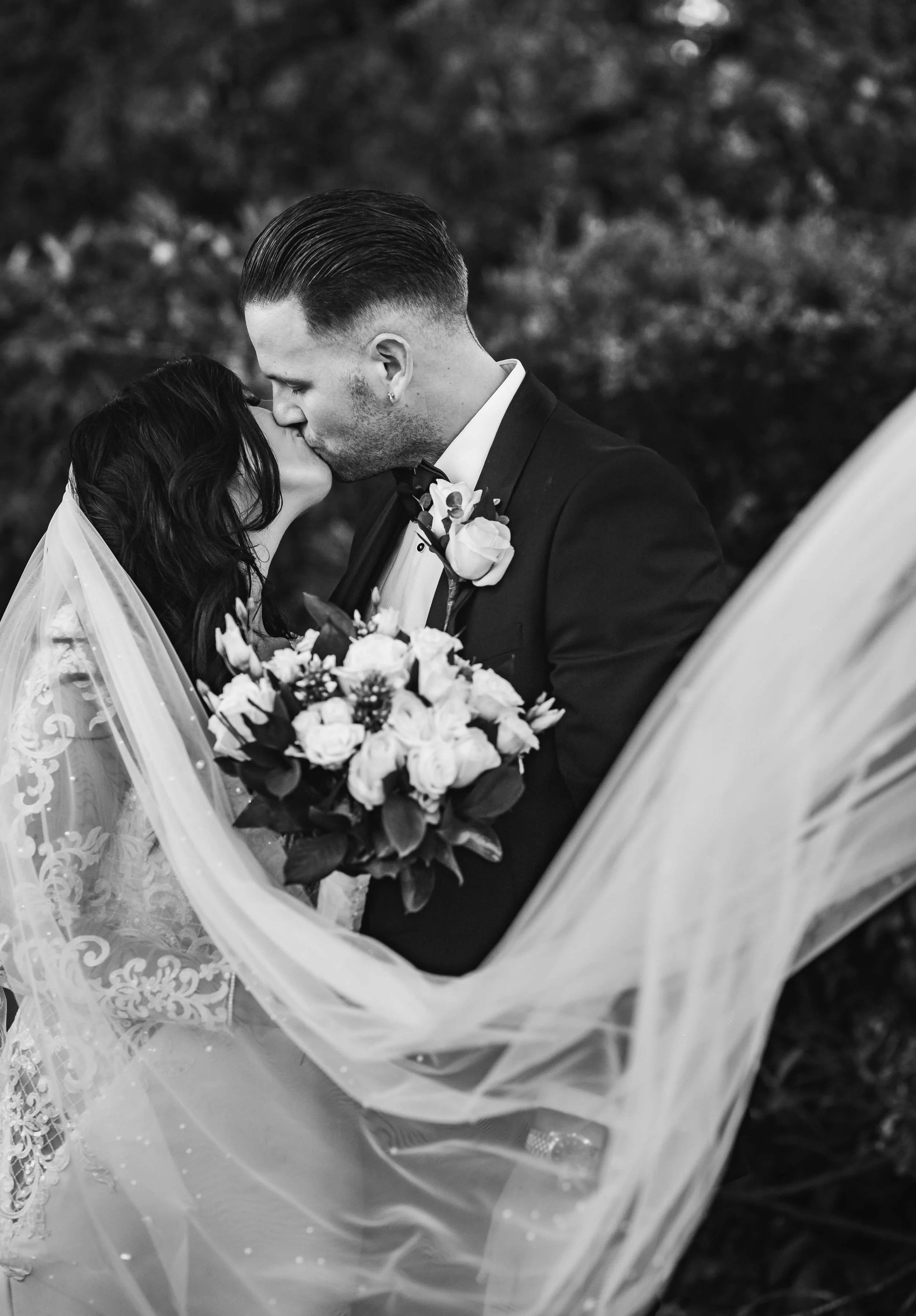 A black and white photo of a bride and groom sharing a kiss, with the bride holding a bouquet of flowers and a wedding veil, outdoors surrounded by foliage.