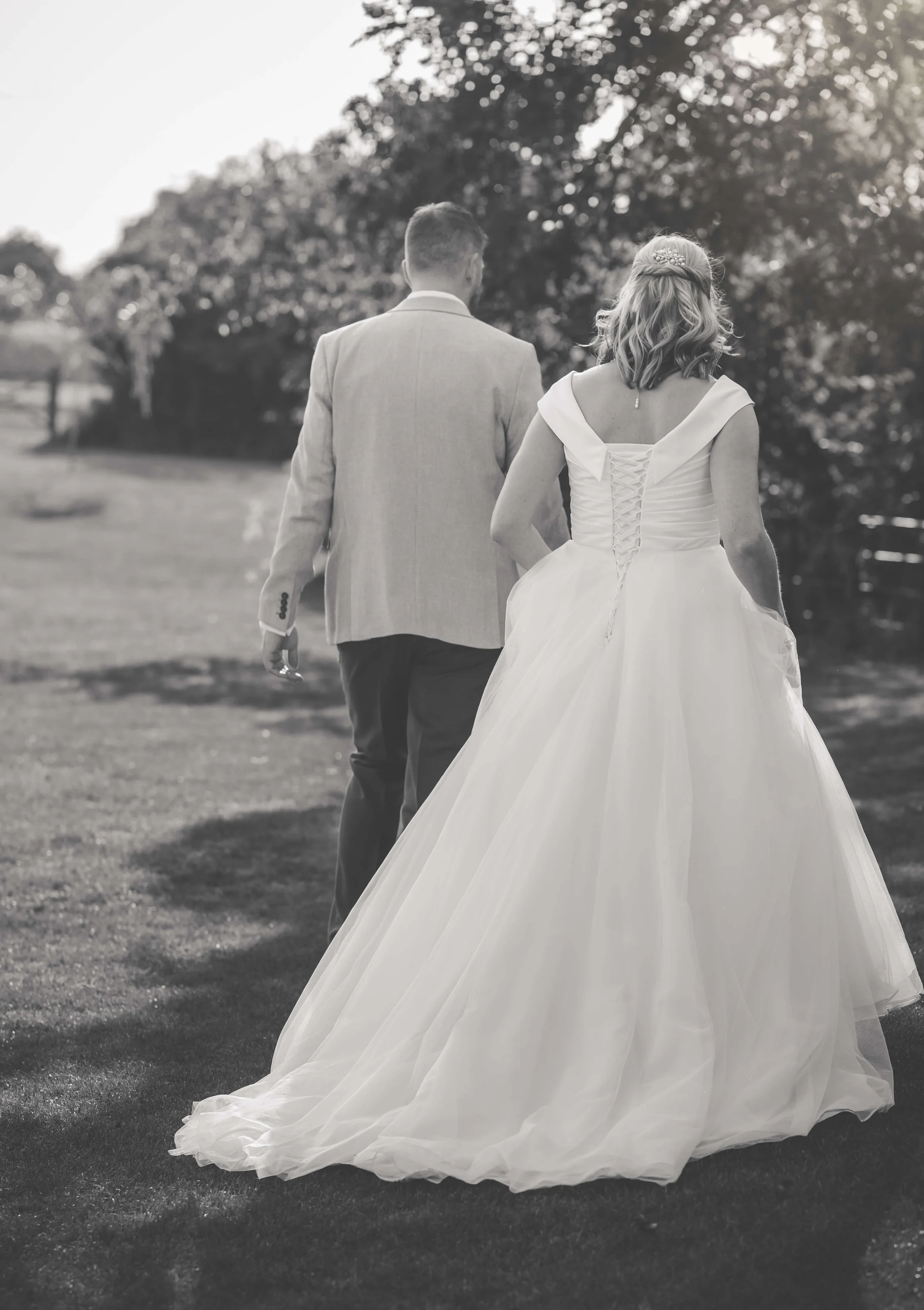 A bride and groom walking away outdoors, the bride in a white wedding gown and the groom in a light suit, with trees and sky in the background.