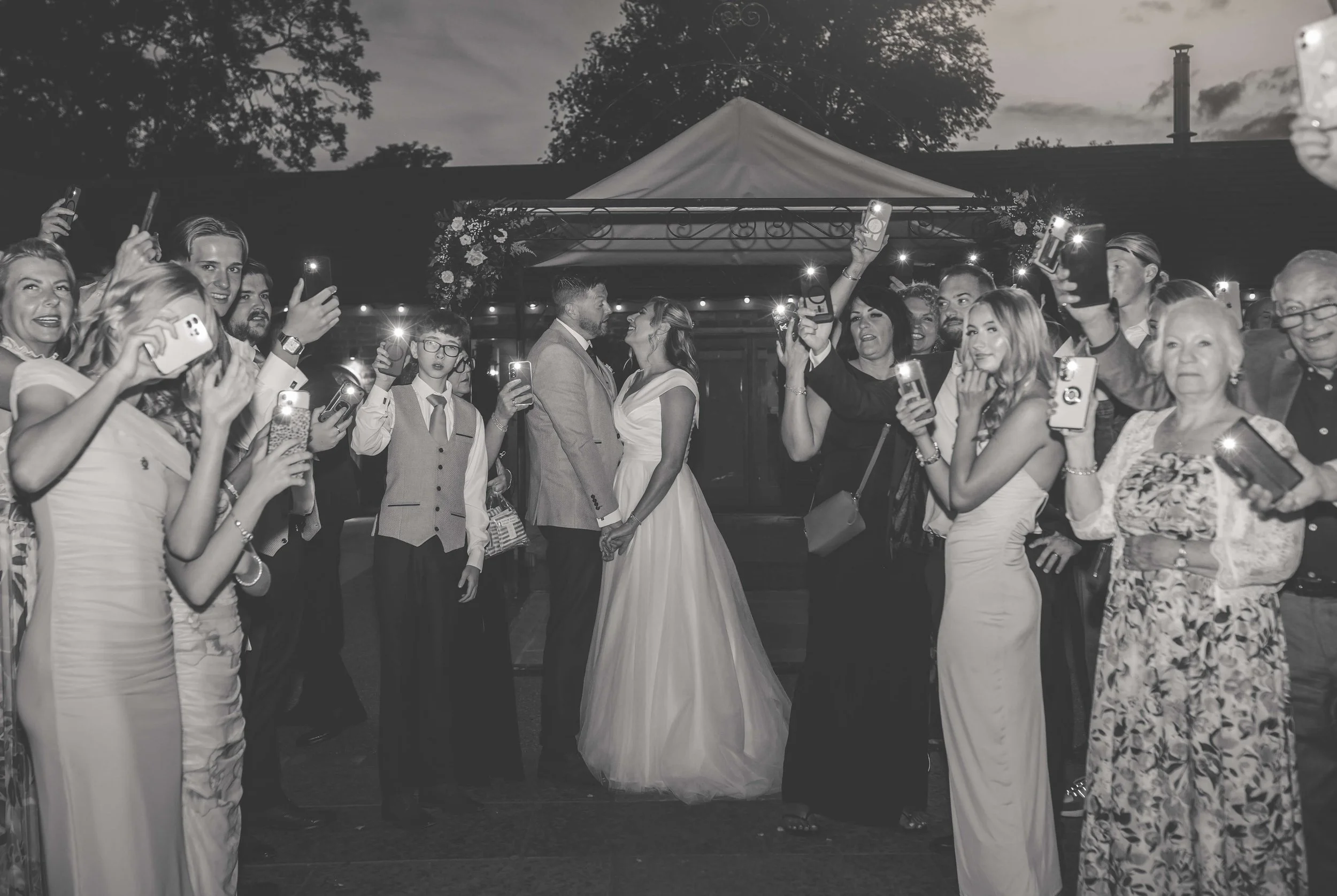 A black-and-white photo of a wedding reception with a bride and groom holding hands and kissing under a canopy, surrounded by guests taking photos with their smartphones.