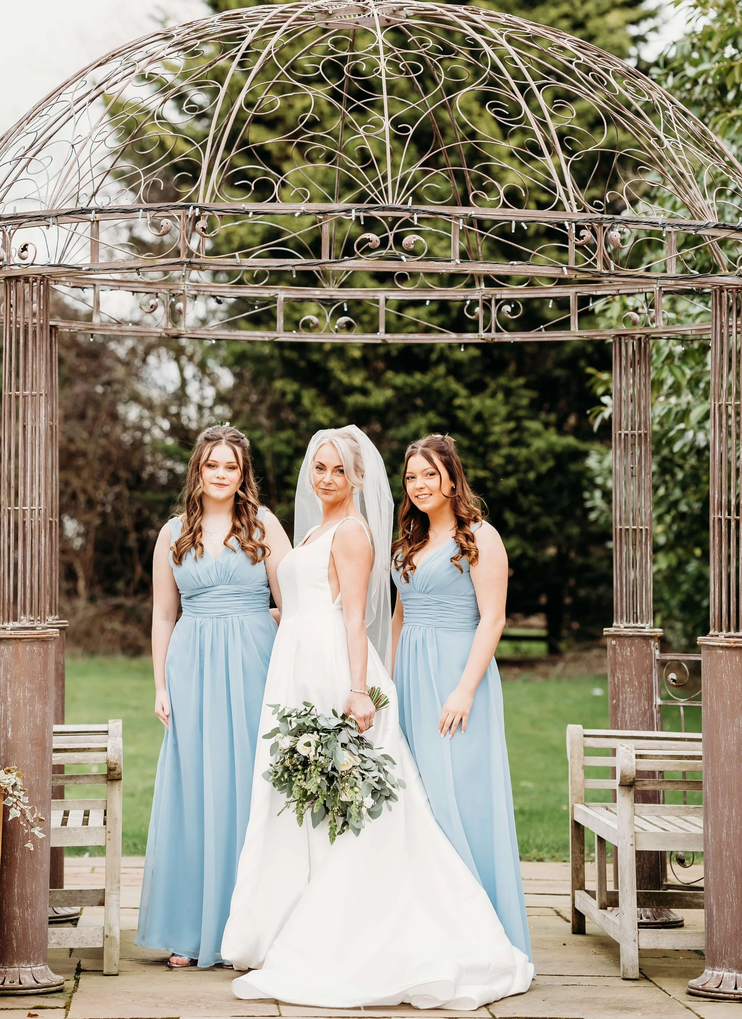 A bride in a white wedding gown holding a bouquet poses with two bridesmaids in light blue dresses under a decorative iron gazebo outdoors.