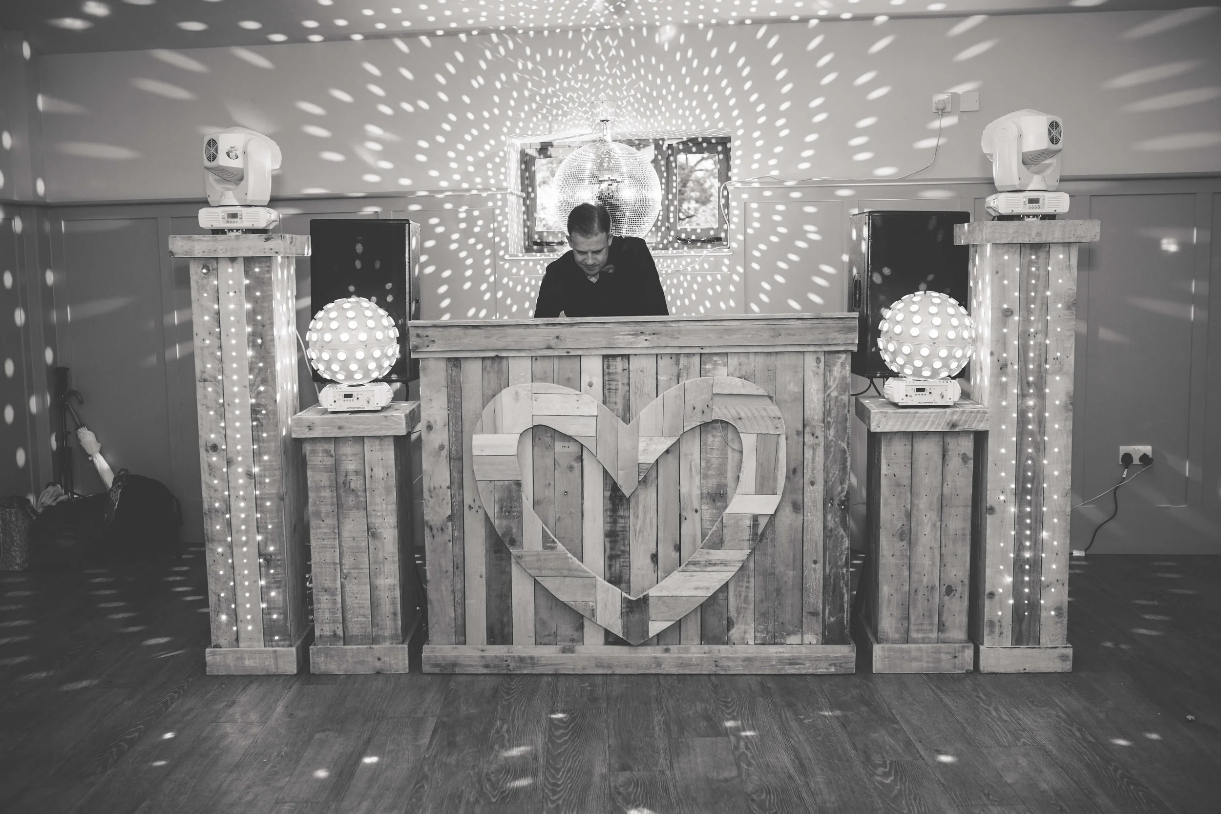 A man DJing at a rustic, wooden wedding reception setup with decorative lights and a disco ball overhead.