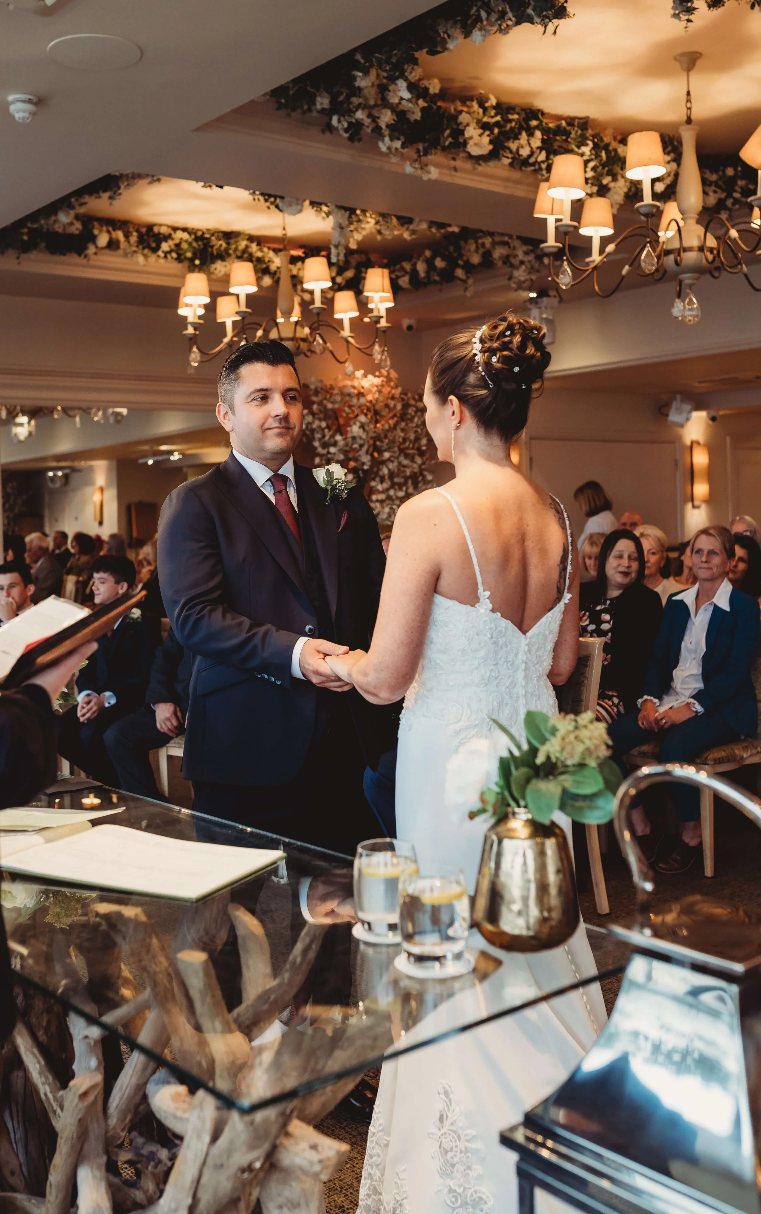 A couple exchanging vows during a wedding ceremony inside a decorated venue, with guests seated in the background, floral arrangements, and warm lighting.