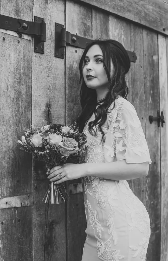 Black and white photo of a woman in a lace dress holding a bouquet of flowers, standing in front of a rustic wooden barn door.