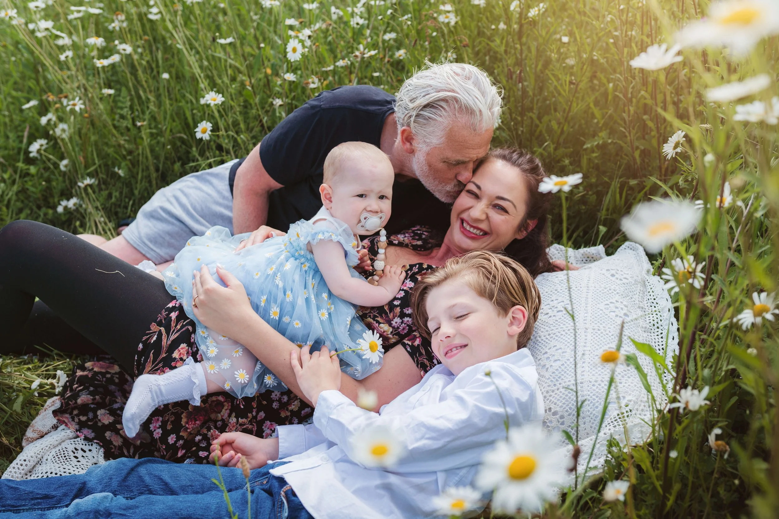 A multigenerational family lying in a field of daisies, shared joyful moment, in a natural outdoor setting.