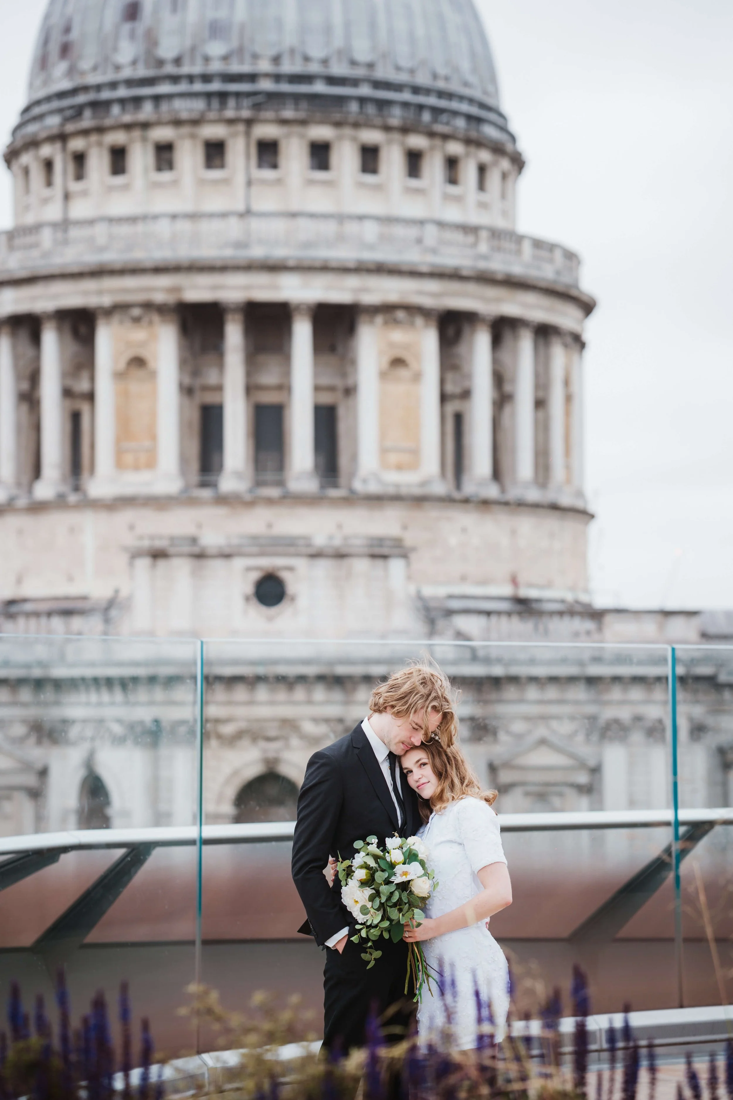 A newlywed couple shares an intimate moment outdoors, holding a bouquet of white flowers in front of a glass barrier. The groom is dressed in a black suit, and the bride is wearing a white lace wedding dress. Behind them is an iconic historic buildin
