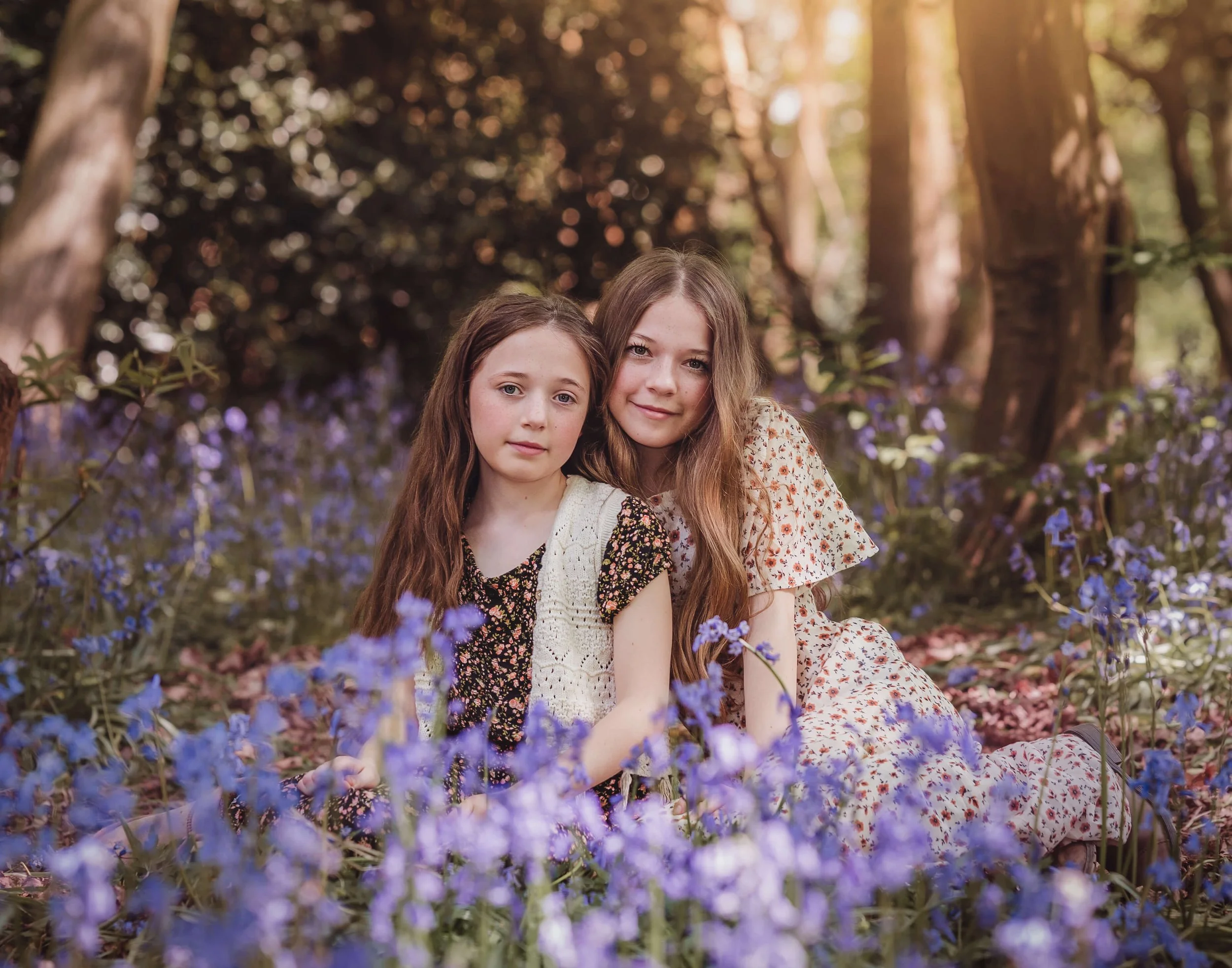 Two young girls sitting amongst purple flowers in a sunlit forest, smiling softly at the camera.