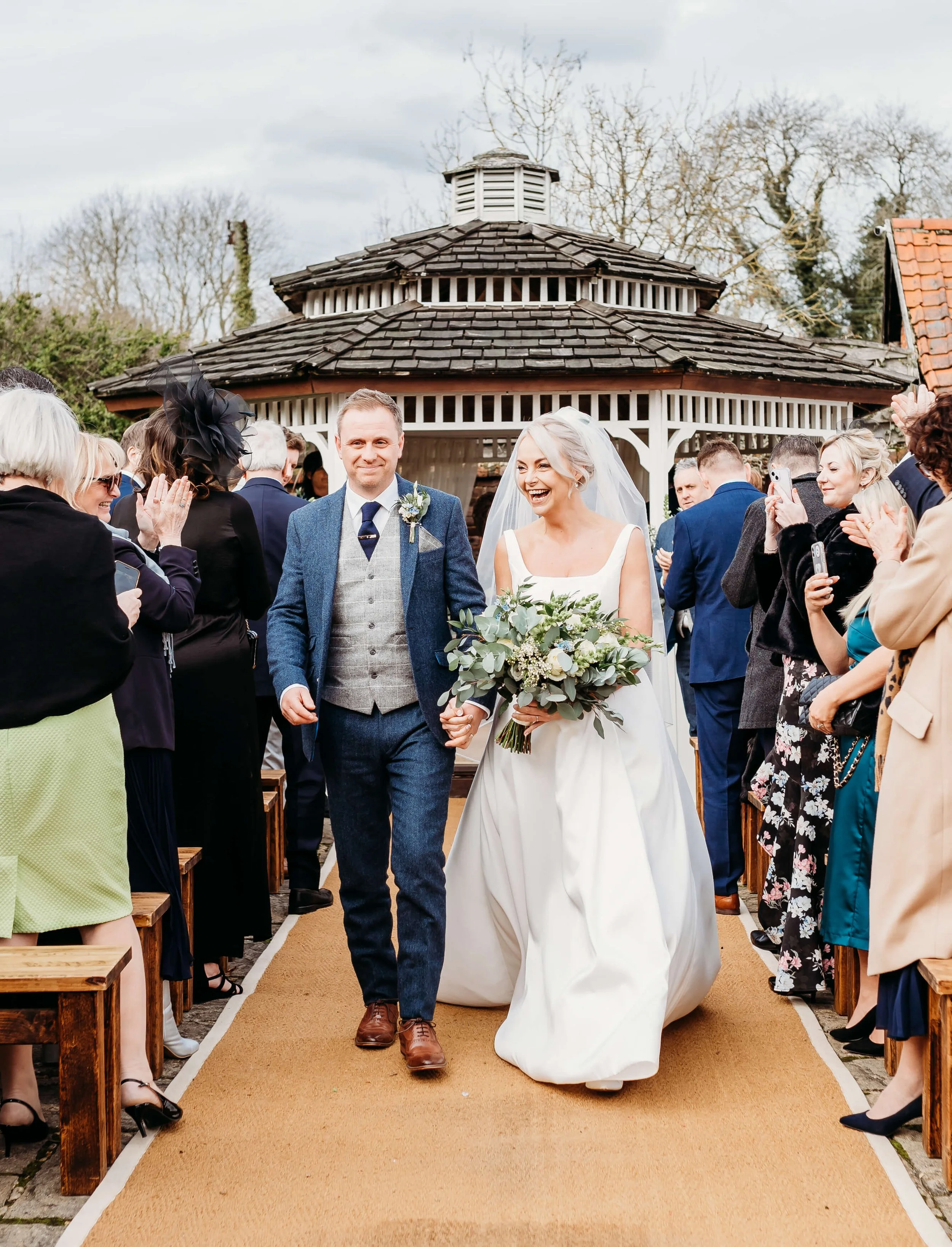 A bride and groom walking down an outdoor aisle, holding hands, surrounded by applauding guests, with a gazebo in the background.