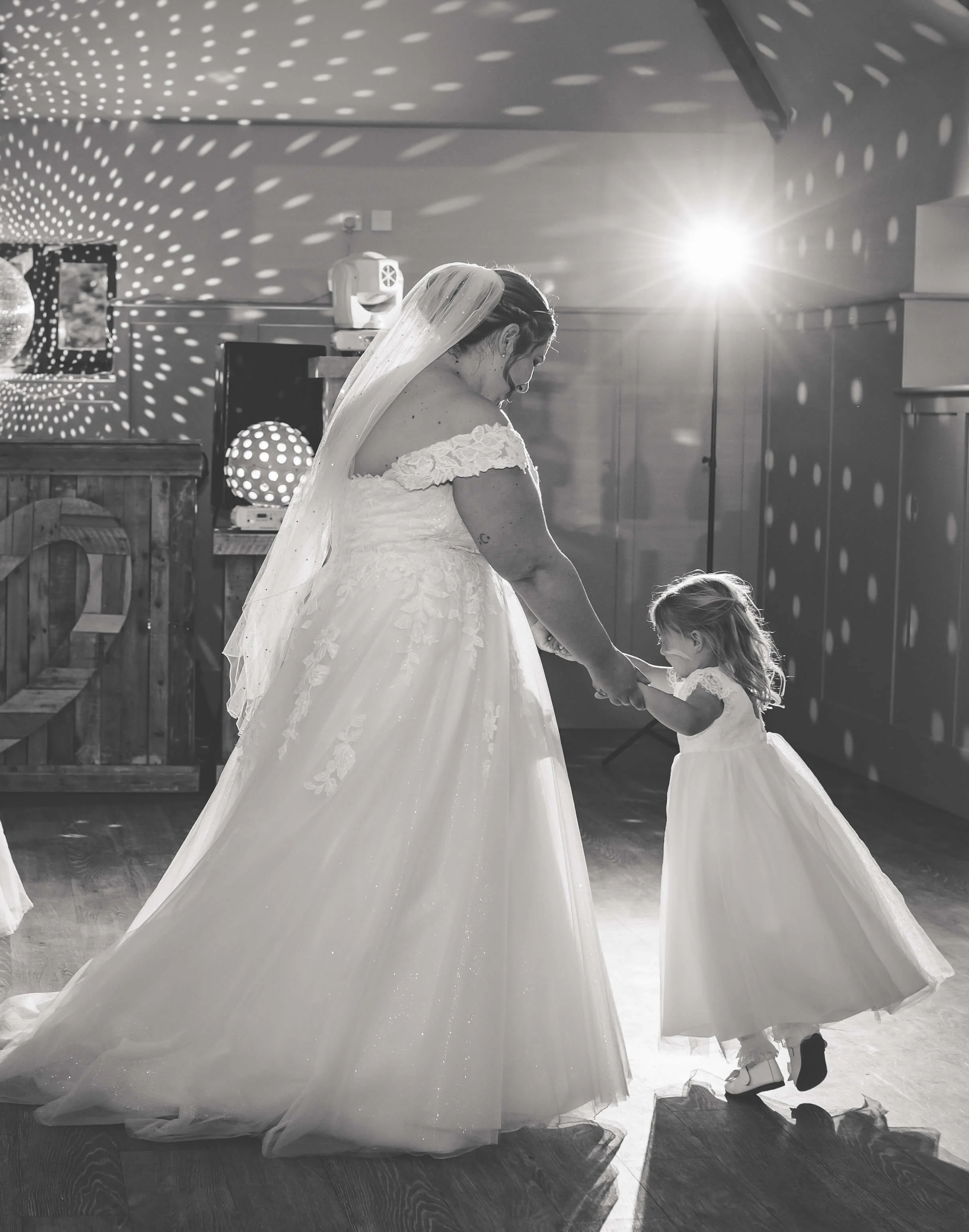 A bride and a young girl dance together in a wedding reception hall, with a bright light shining in the background creating a pattern of light and shadow.